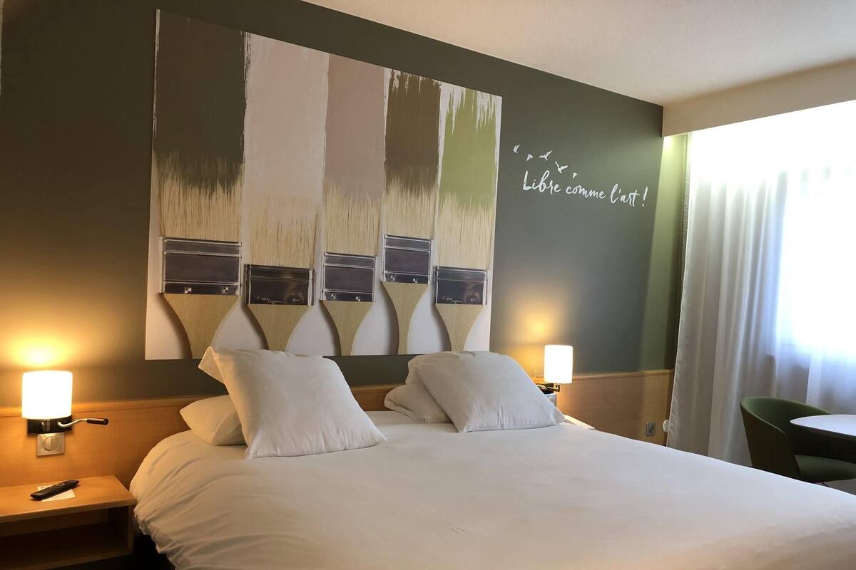 A hotel room with a large bed, two bedside lamps, a green chair, and a wall with a painting and text.