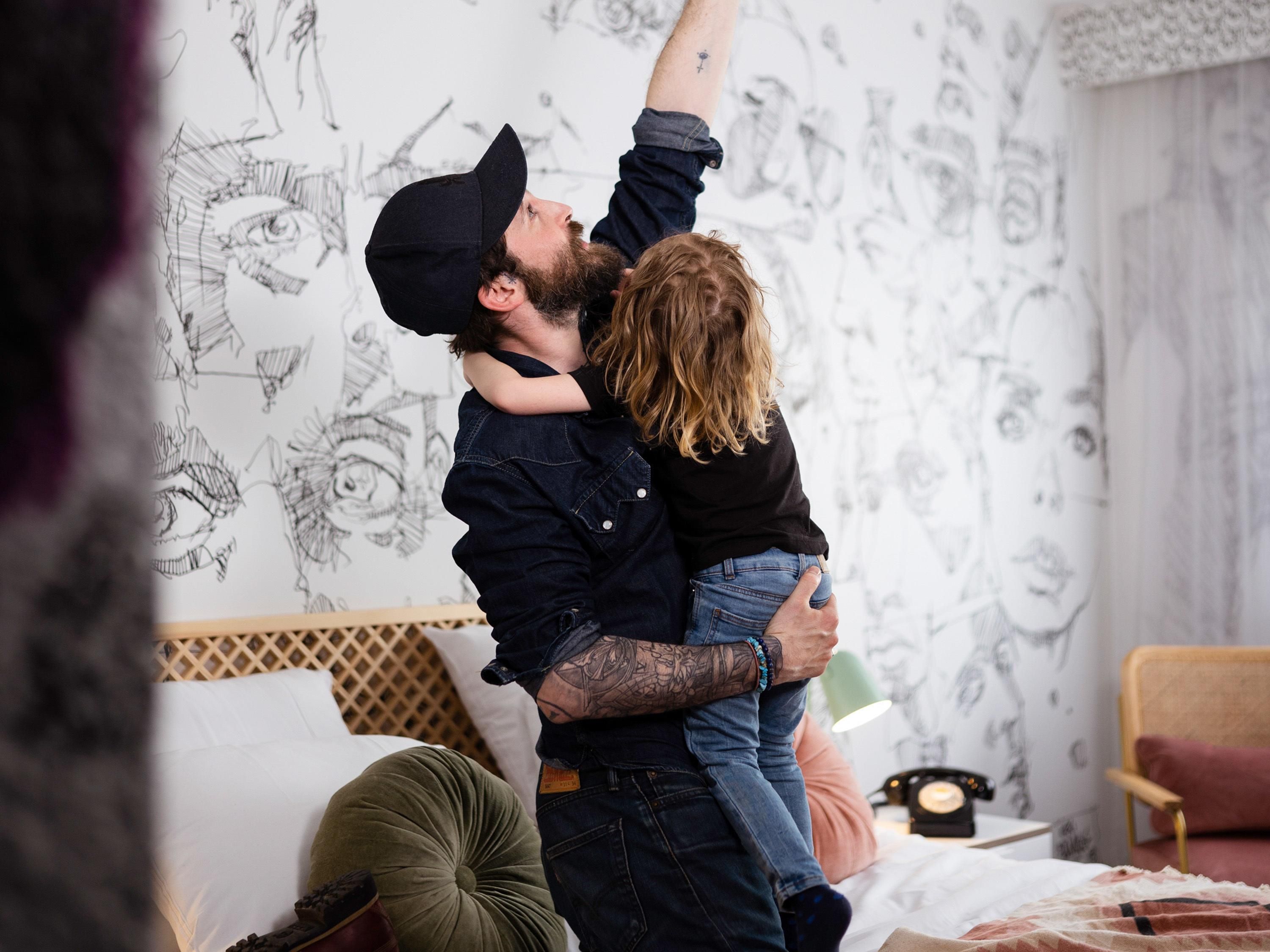 A man lifting a child in a room with a bed, a wooden headboard, and a wall with sketches.