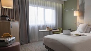 A hotel room with a bed, a desk, a chair, a suitcase, and a window with curtains. - Mercure Reims Parc des Expositions (Reims)