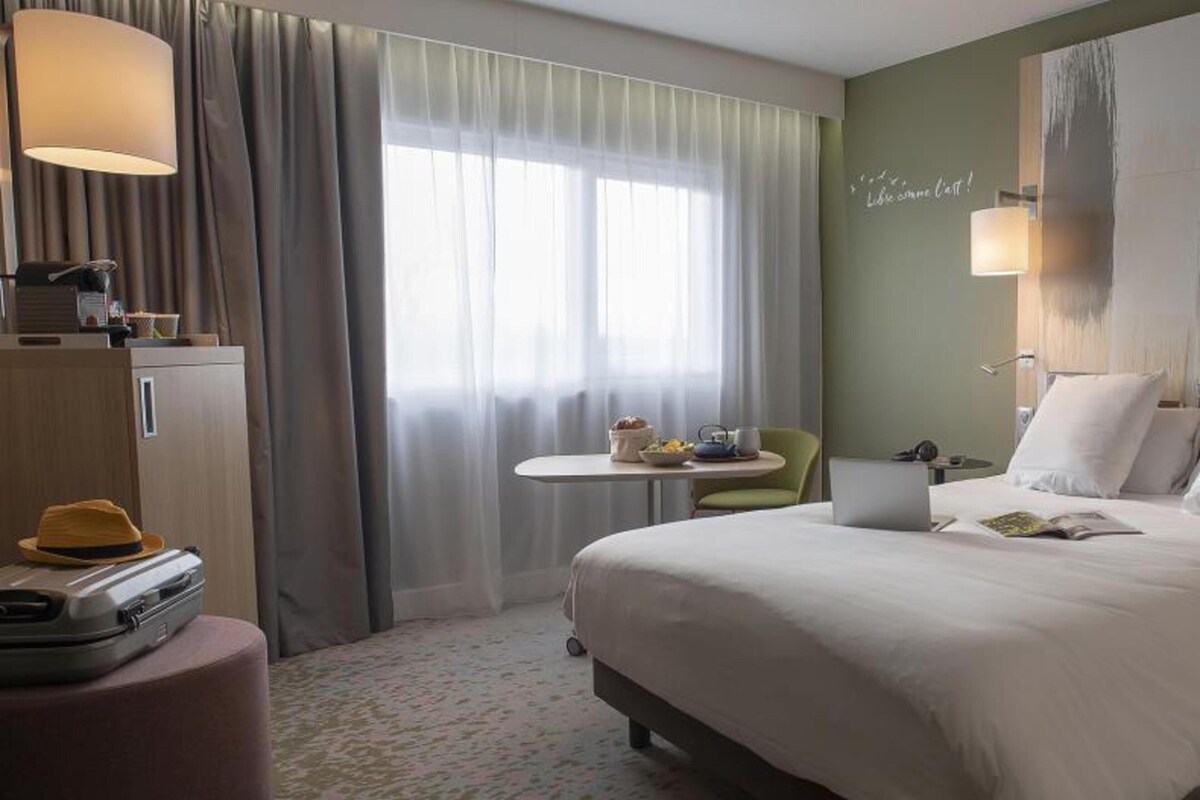 A hotel room with a bed, a desk, a chair, a suitcase, and a window with curtains.