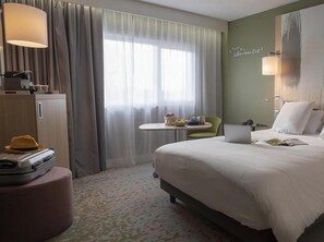 A hotel room with a bed, a desk, a chair, a suitcase, and a window with curtains. - Mercure Reims Parc des Expositions (Reims)