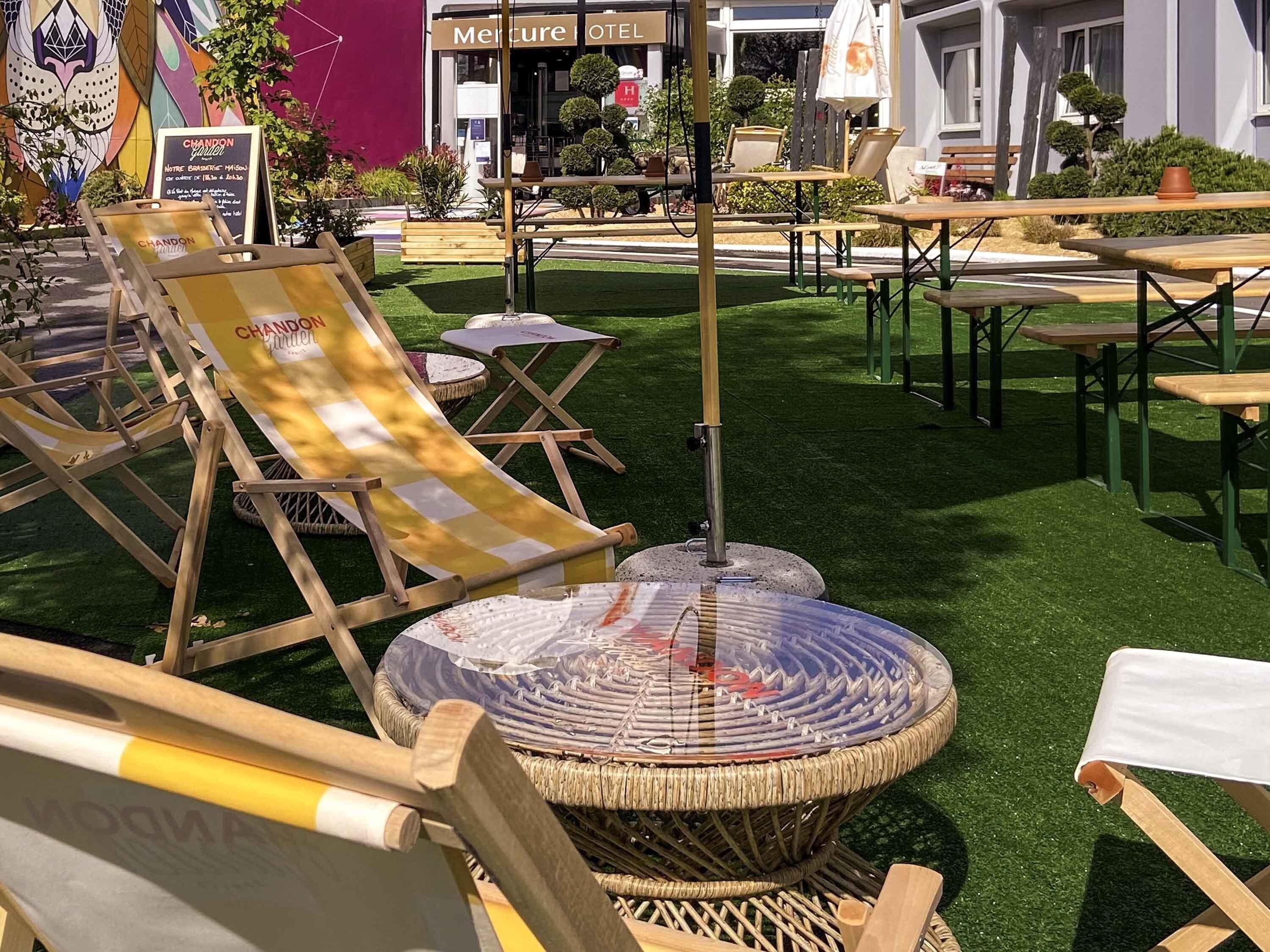 An outdoor seating area with wicker chairs and tables, a round table with a glass top, and a sign for 'Mercure OTEL' in the background.