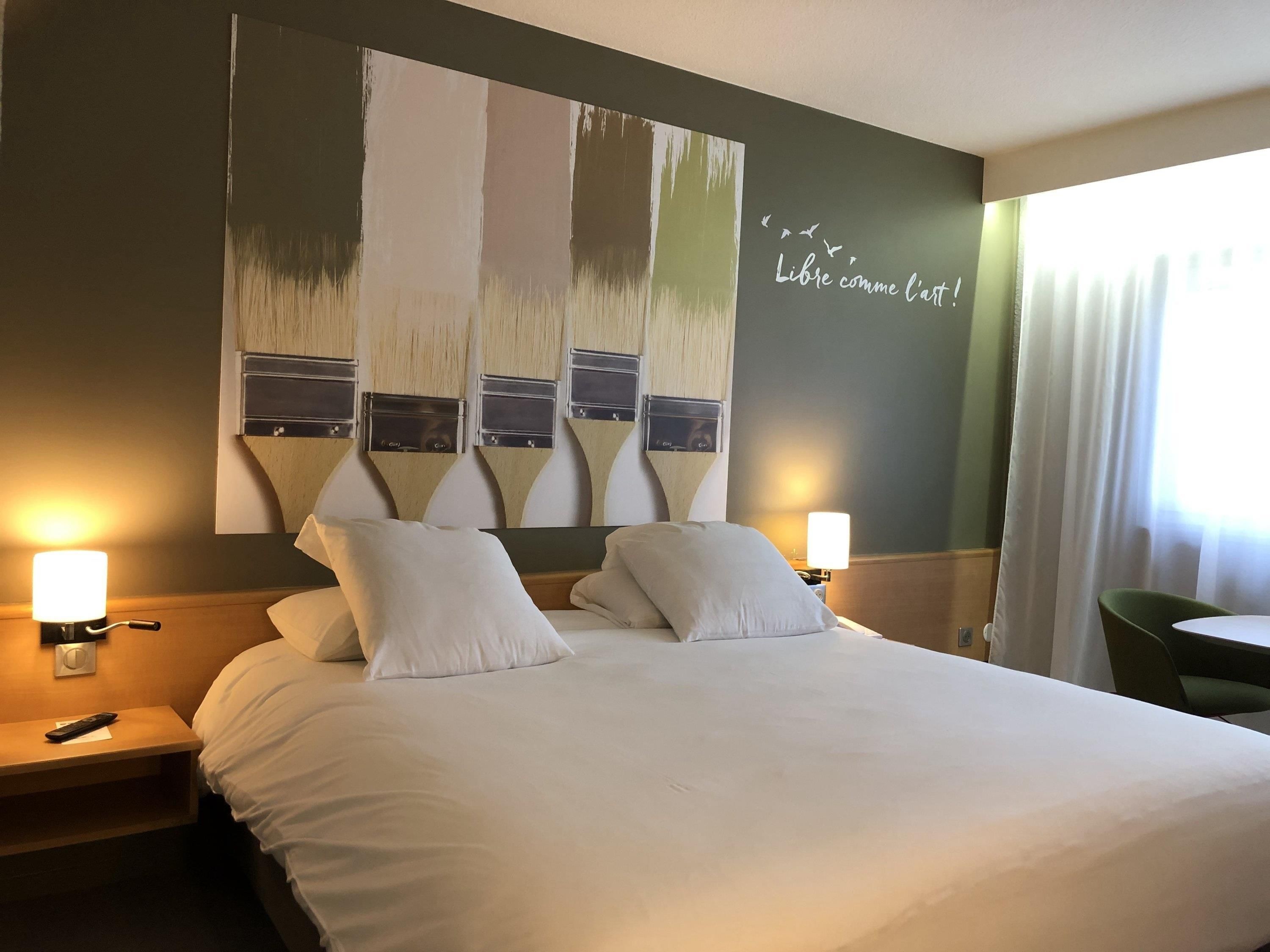 A hotel room with a large bed, two bedside lamps, a green chair, and a wall with a painting and text.