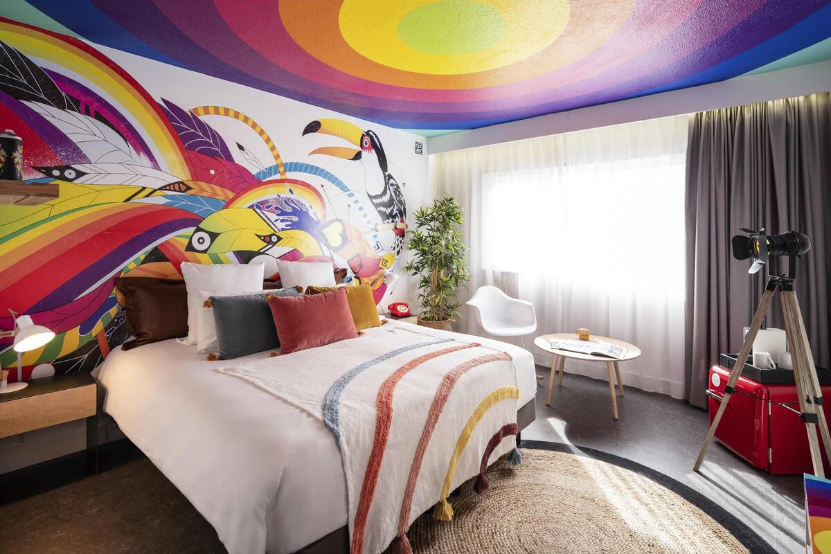 A hotel room with a colorful mural, a bed with striped bedding, a small round table, a tripod, and a potted plant.
