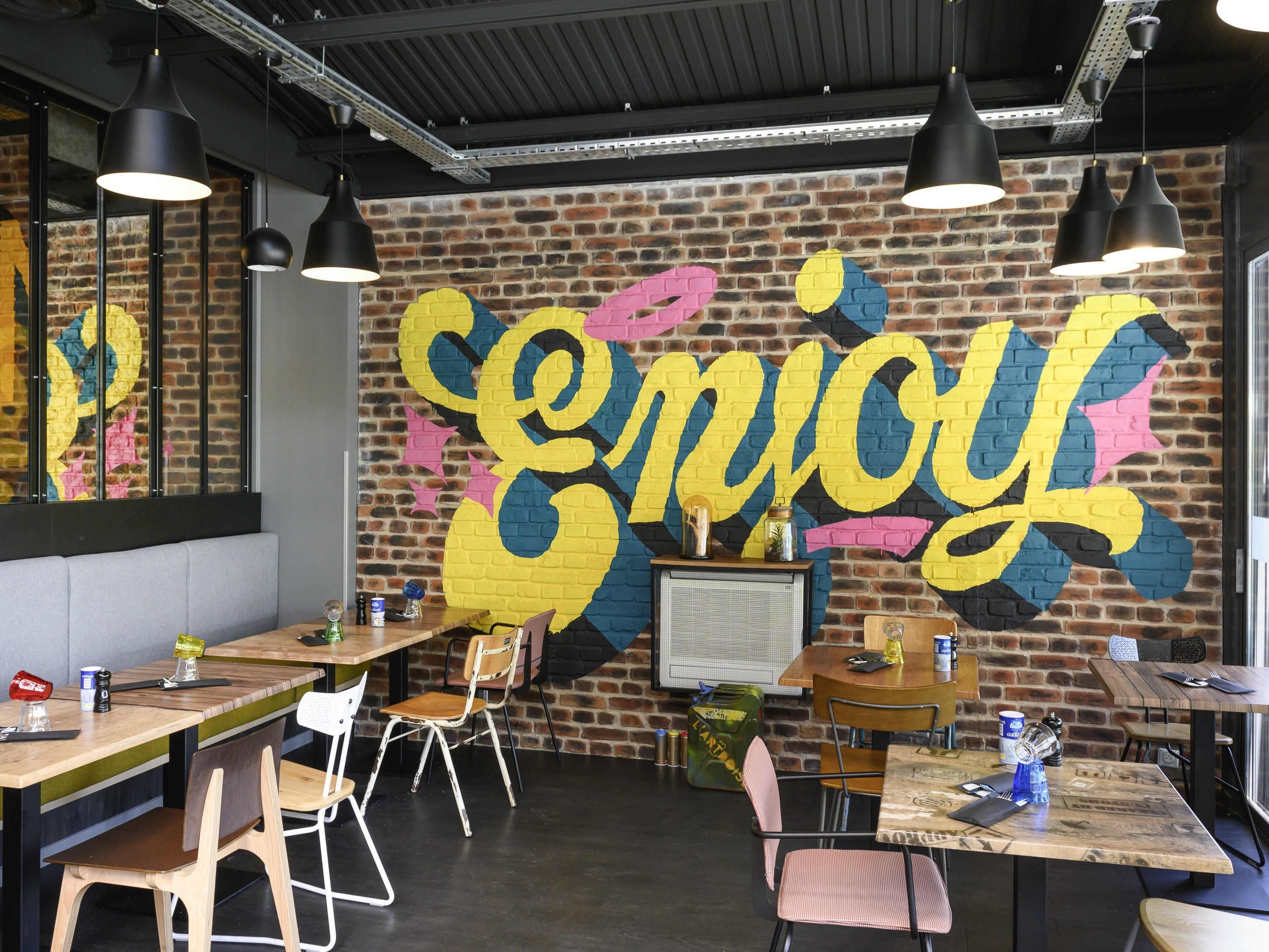 A restaurant interior with brick walls, black pendant lighting, and a large 'enjoy' mural. Tables and chairs are arranged for dining.