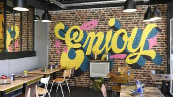 A restaurant interior with brick walls, black pendant lighting, and a large 'enjoy' mural. Tables and chairs are arranged for dining.