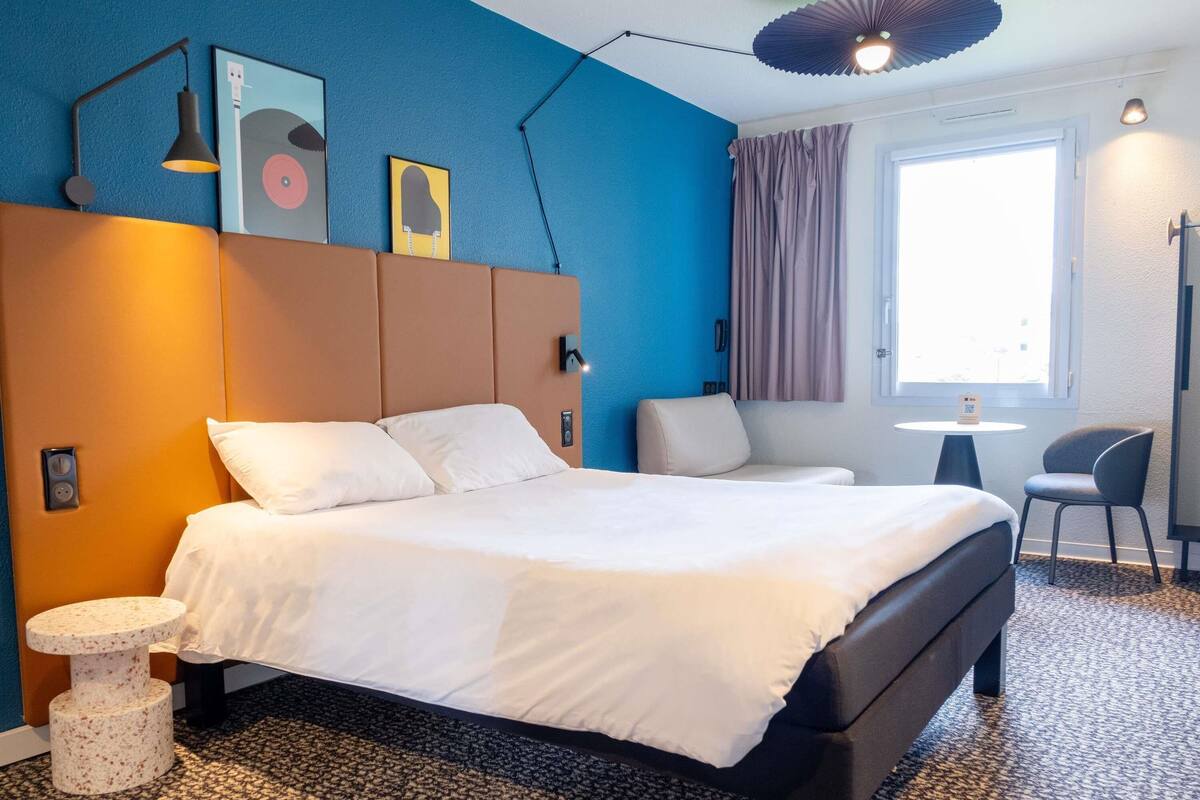 Una habitació d'hotel moderna amb un llit gran, una taula petita, una cadira i una finestra amb cortines.