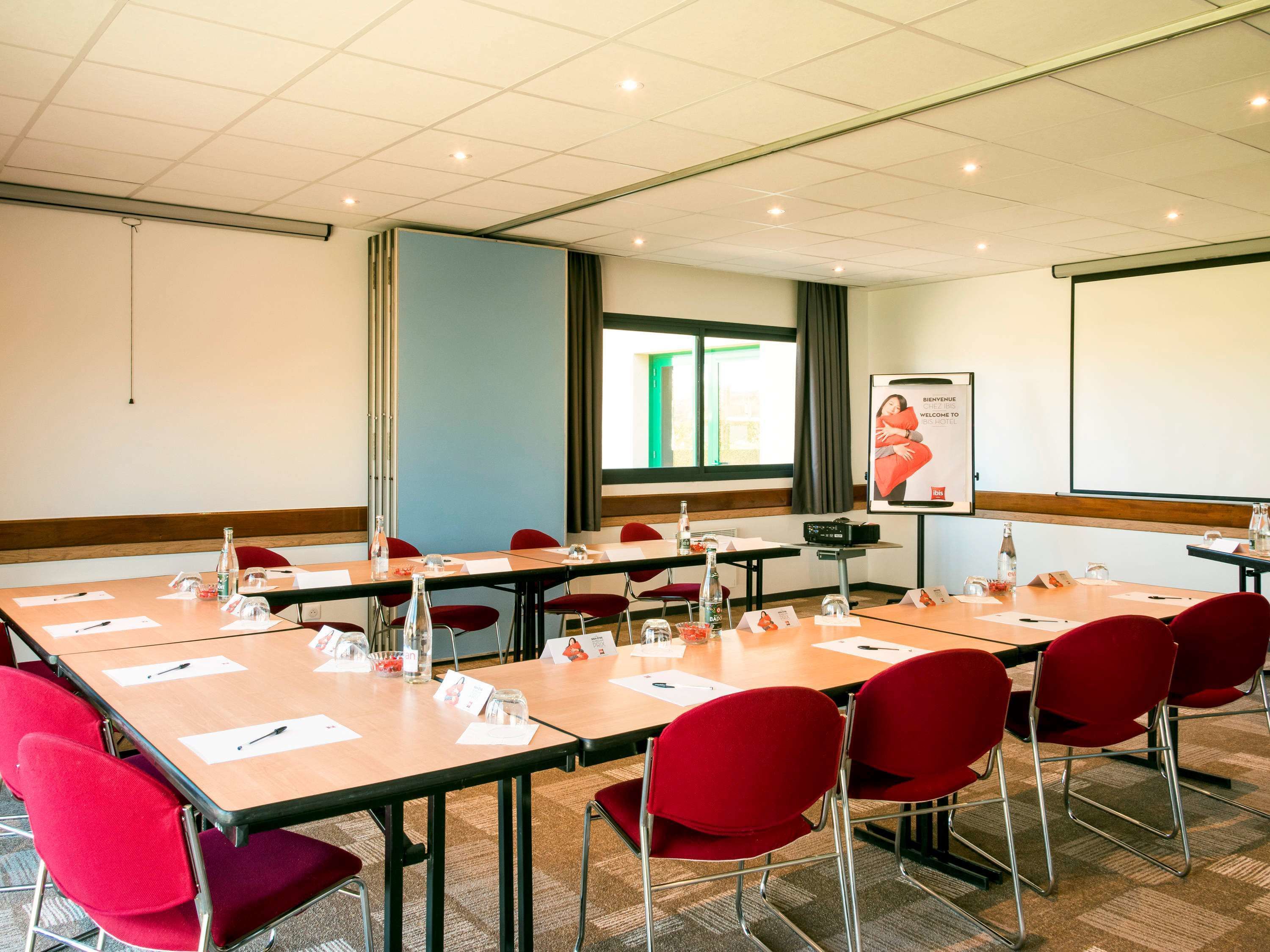 A conference room with multiple tables and red chairs, equipped with nameplates, water bottles, and a projector screen displaying a welcome message.