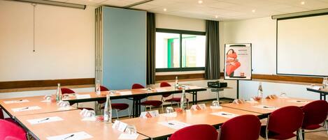 A conference room with multiple tables and red chairs, equipped with nameplates, water bottles, and a projector screen displaying a welcome message.