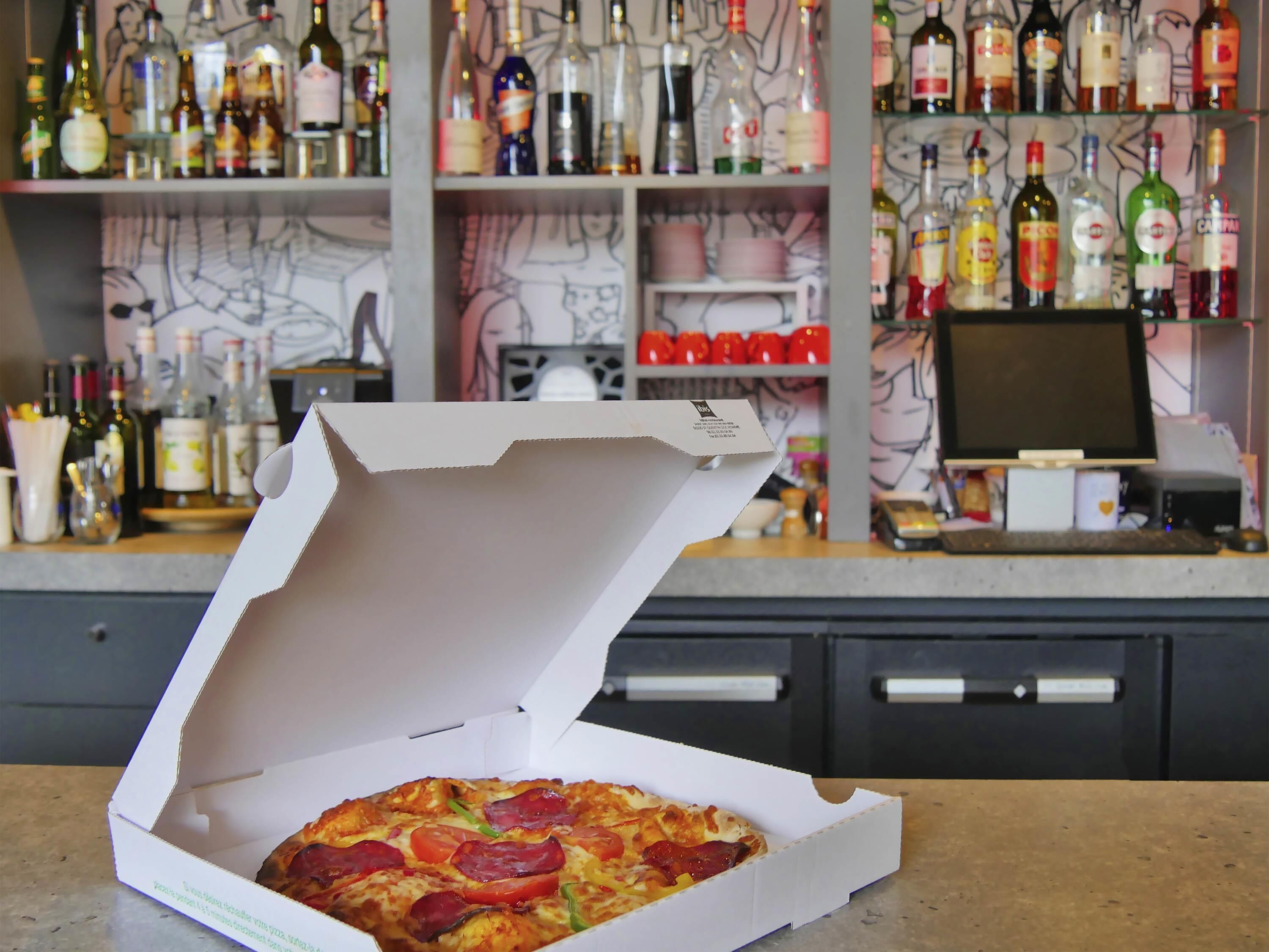 Una pizza en una caja sobre una encimera, con una barra y estantes llenos de botellas al fondo.