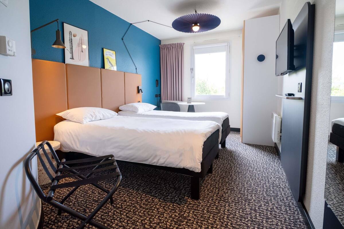 Habitació d'hotel amb dos llits, una taula de treball, una cadira, un televisor i una finestra amb cortines.