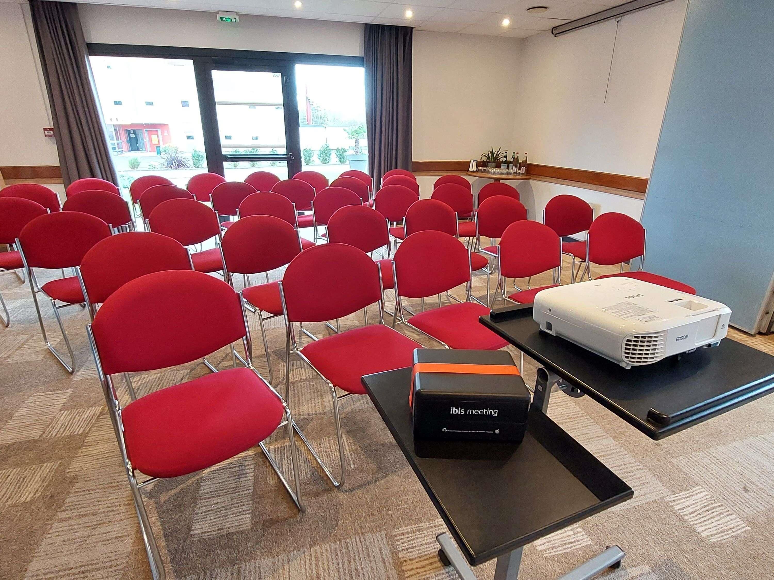 A conference room with rows of red chairs, a projector, and an ibis meeting box on a table.