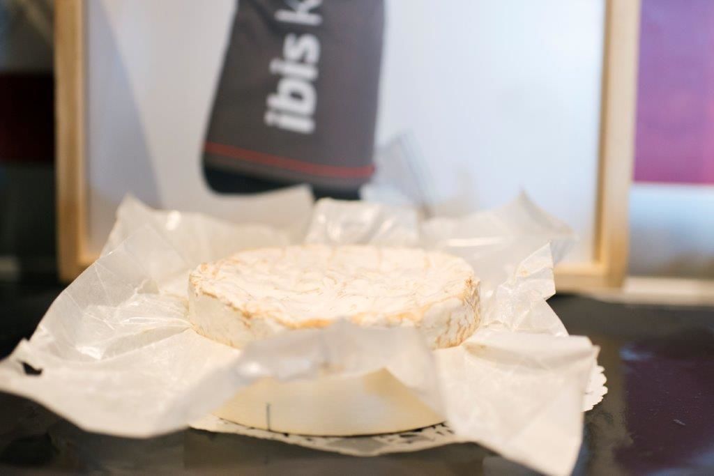 Un queso redondo sobre una mesa, con una bolsa de la marca al fondo.