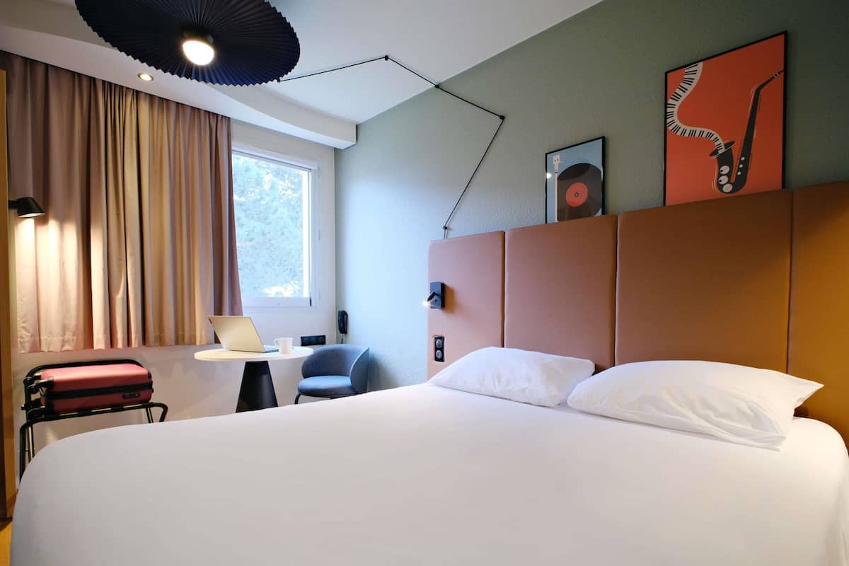 Ein Hotelzimmer mit einem großen Bett, einem Schreibtisch mit Laptop, einem Stuhl, einem Telefon und einem Fenster mit Vorhängen.