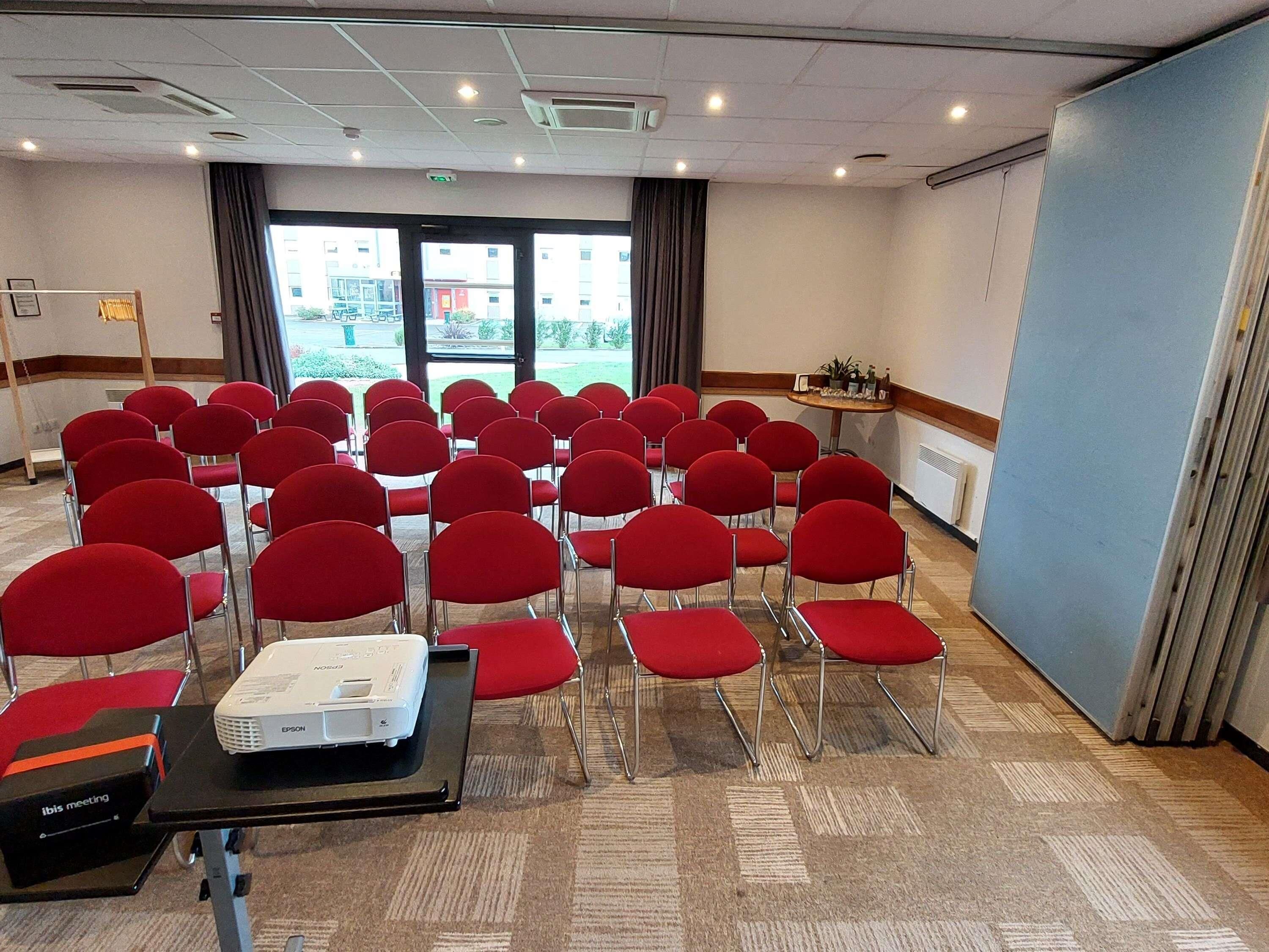Een vergaderzaal met rijen rode stoelen, een beamer op een tafel en uitzicht buiten door grote ramen.