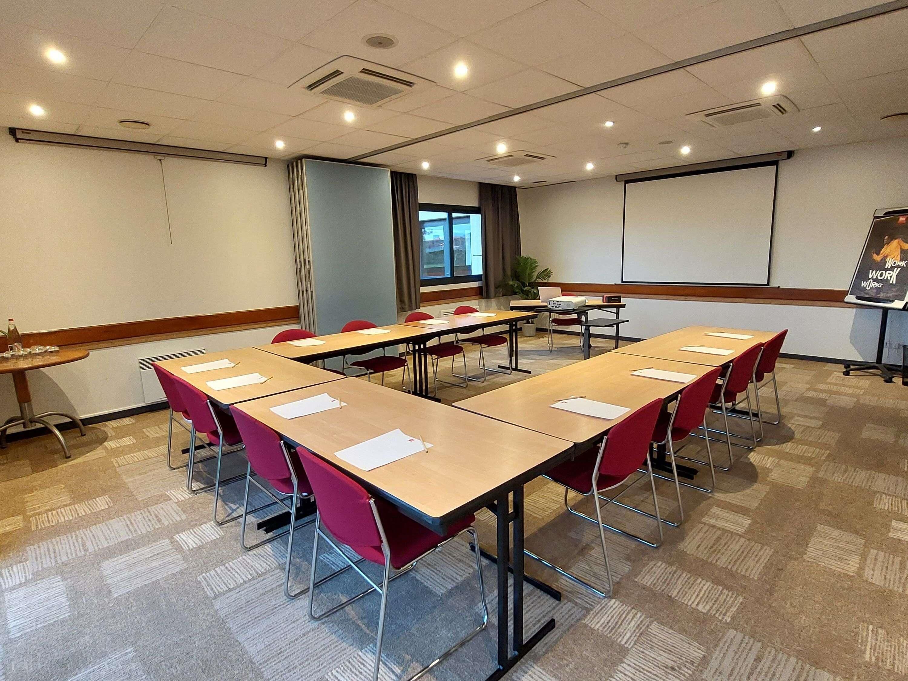 A conference room with long tables, red chairs, a projector screen, and a presentation screen.