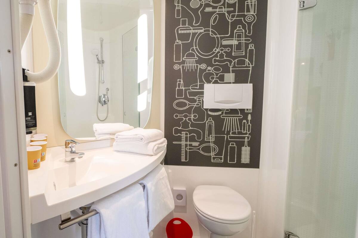 Un baño moderno con un lavamanos blanco, un espejo y un inodoro. Cuenta con una ducha con manguera, un cubo de basura rojo y una pared negra con iconos de baño en blanco.