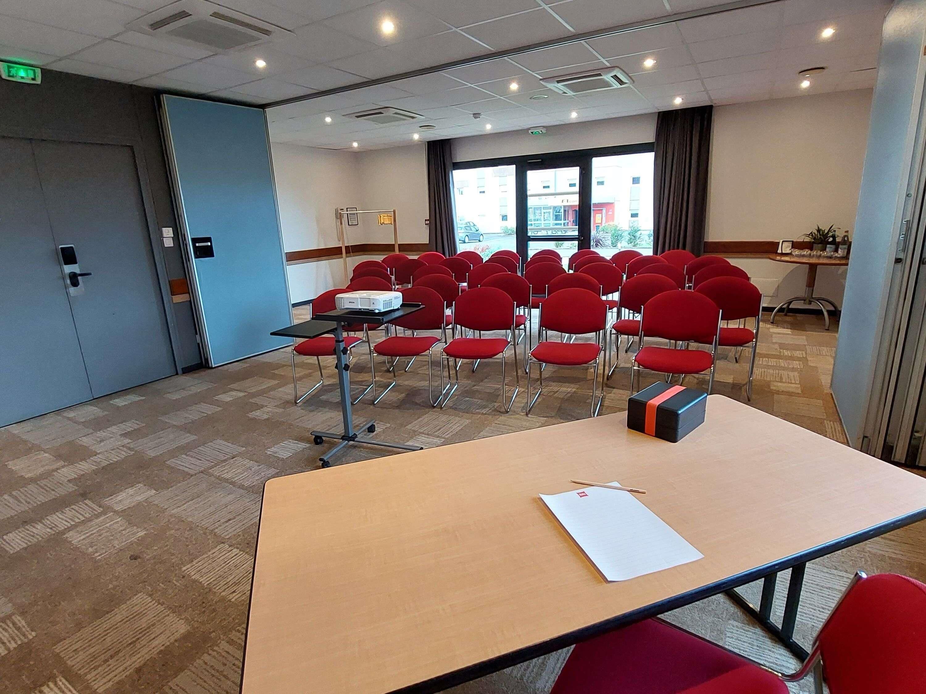 A conference room with a large table, red chairs, a projector, and a window with a view of buildings outside.
