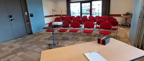 A conference room with a large table, red chairs, a projector, and a window with a view of buildings outside.