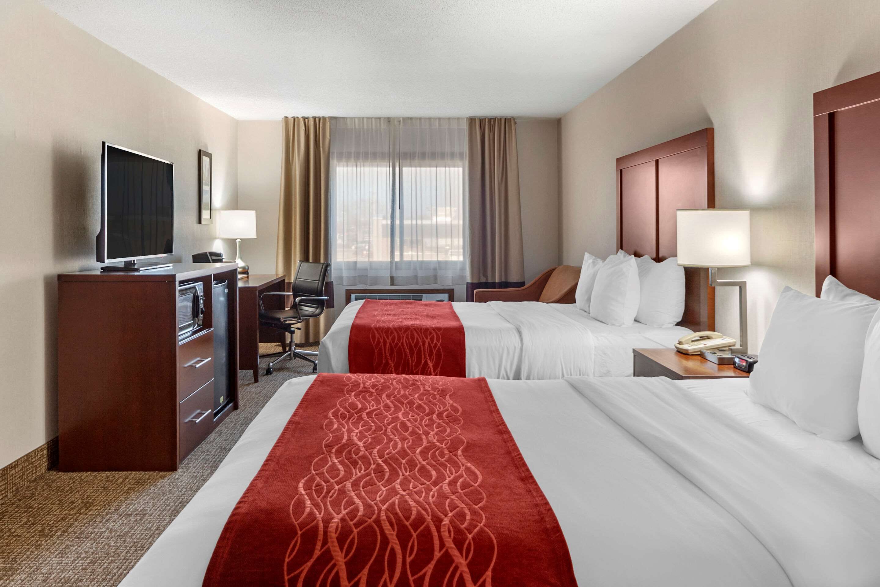 Quarto de hotel com duas camas, uma escrivaninha com televisão, uma cadeira e uma luminária de mesa.