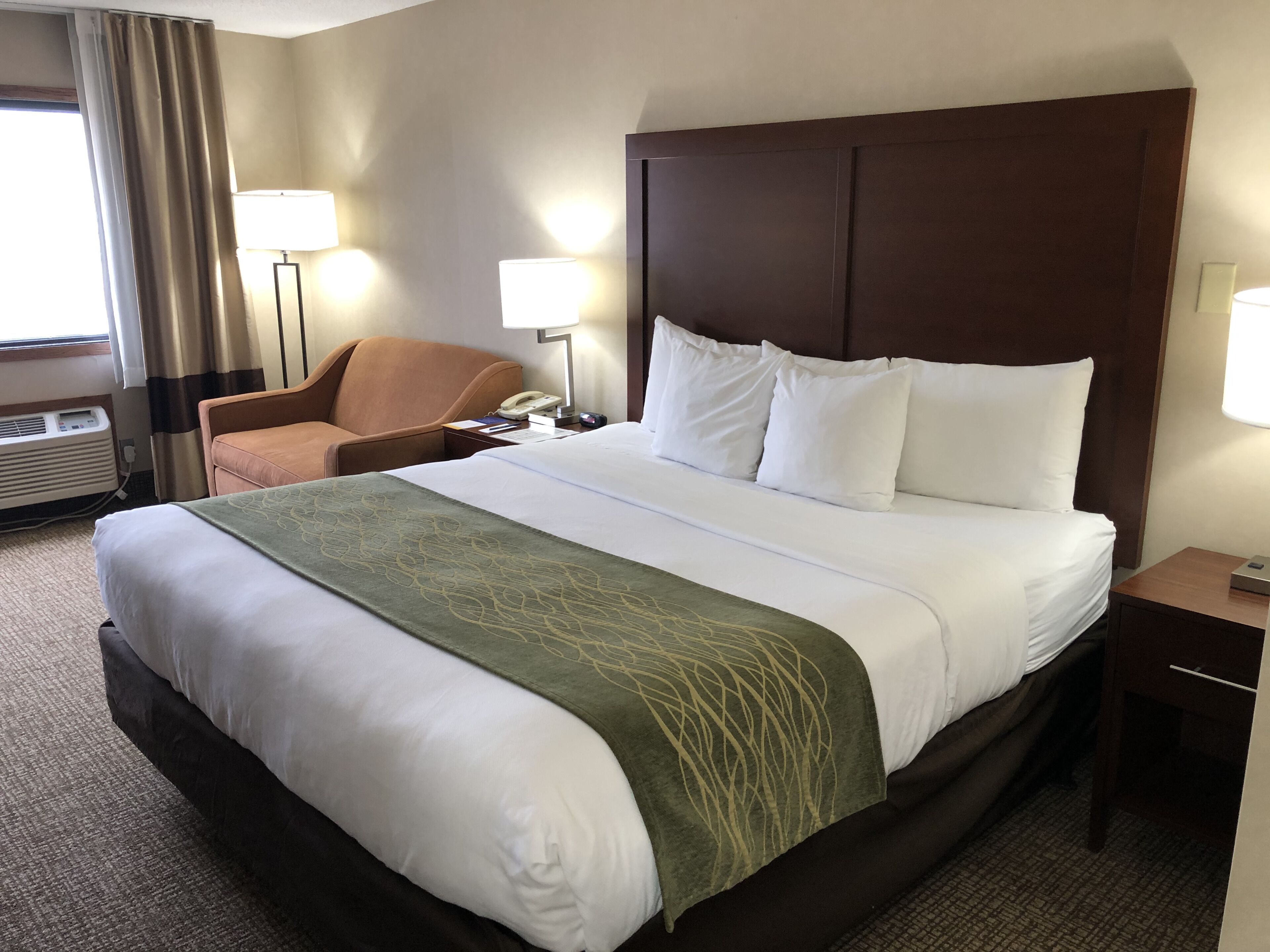 Una habitación de hotel con una cama grande, cabecera, dos lámparas de noche, una silla, un escritorio con teléfono y una ventana con cortinas.