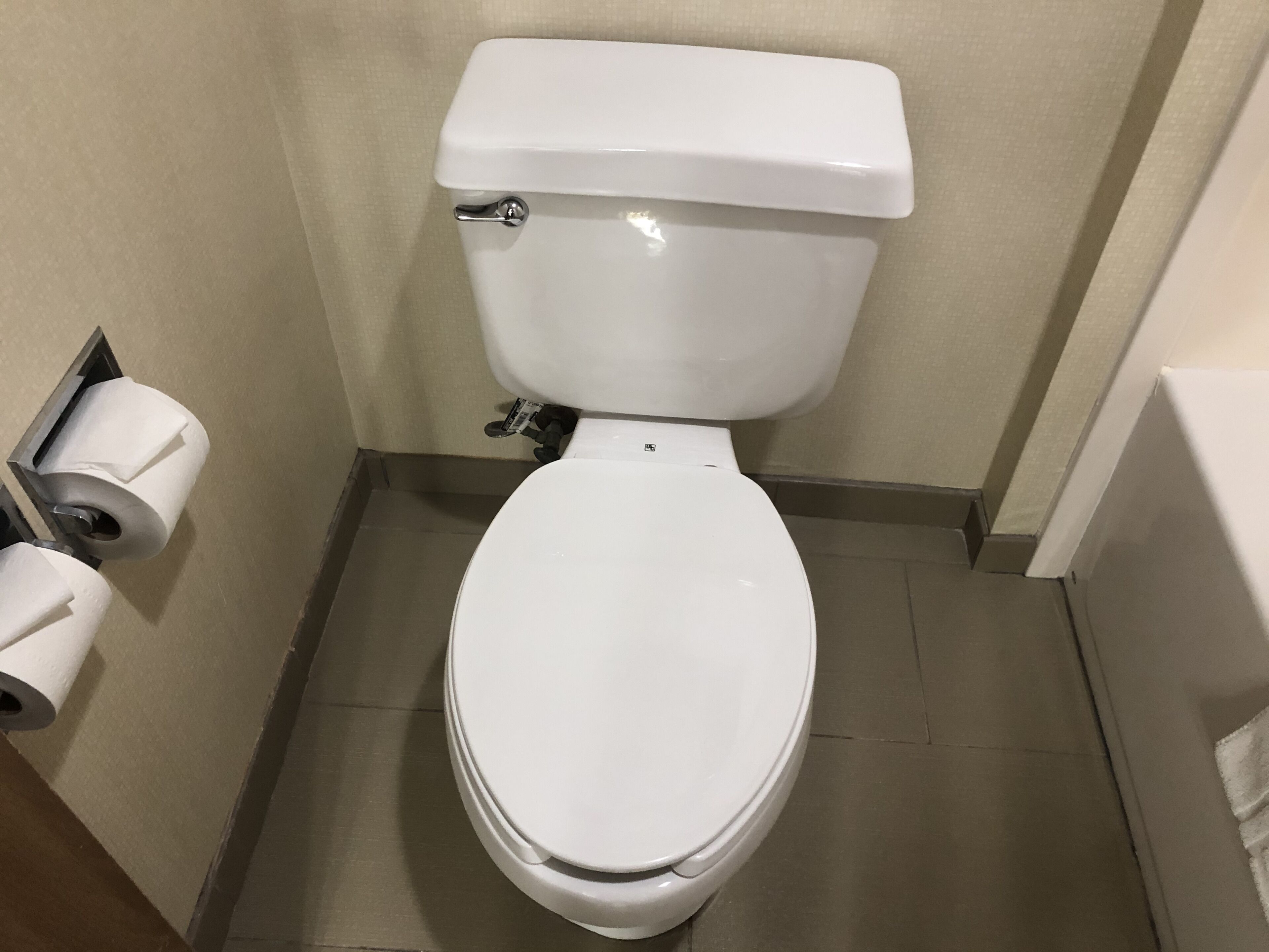 A white toilet with a tank and a flush handle, located in a small bathroom with beige walls and a tiled floor.