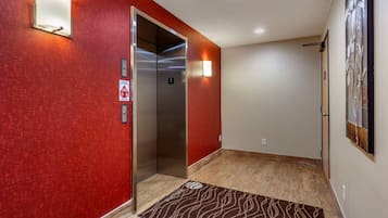 Un pasillo con pared roja, un ascensor, un felpudo y una lámpara de pared.
