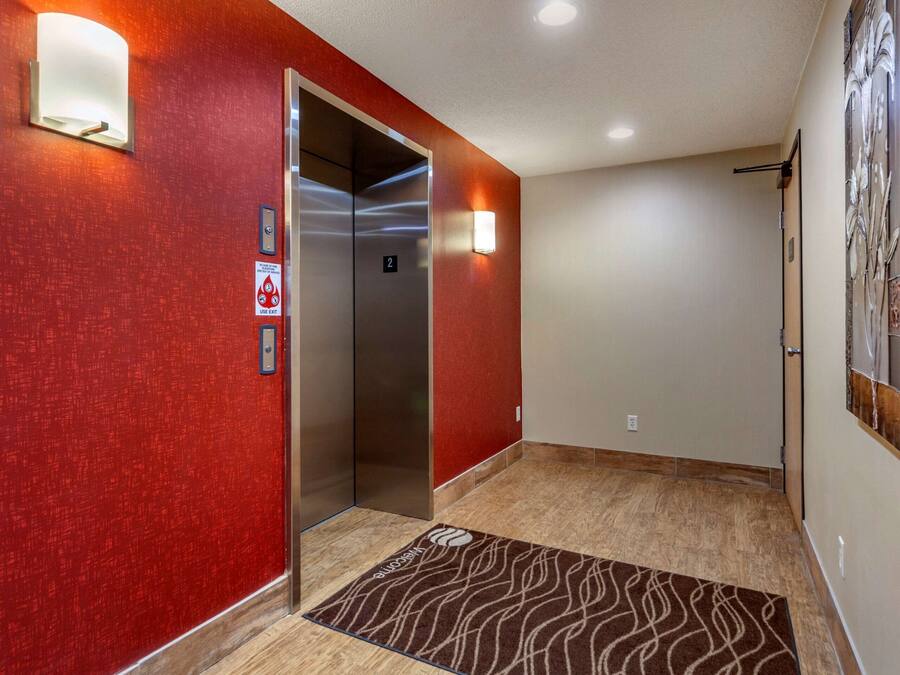 Un pasillo con pared roja, un ascensor, un felpudo y una lámpara de pared.