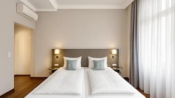 A hotel room with a large bed, two bedside tables with lamps, an air conditioning unit, and a view of the outside through sheer curtains.