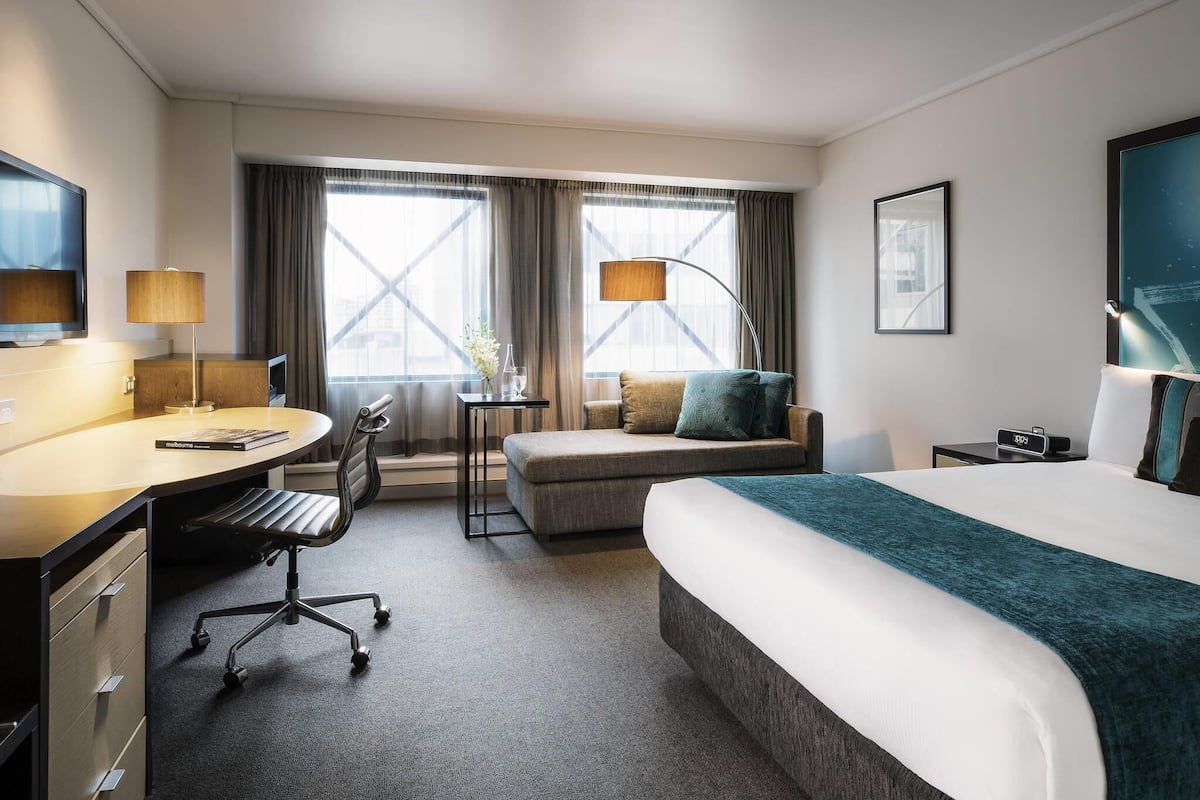 Habitación de hotel con cama, escritorio, silla y dos ventanas con cortinas.