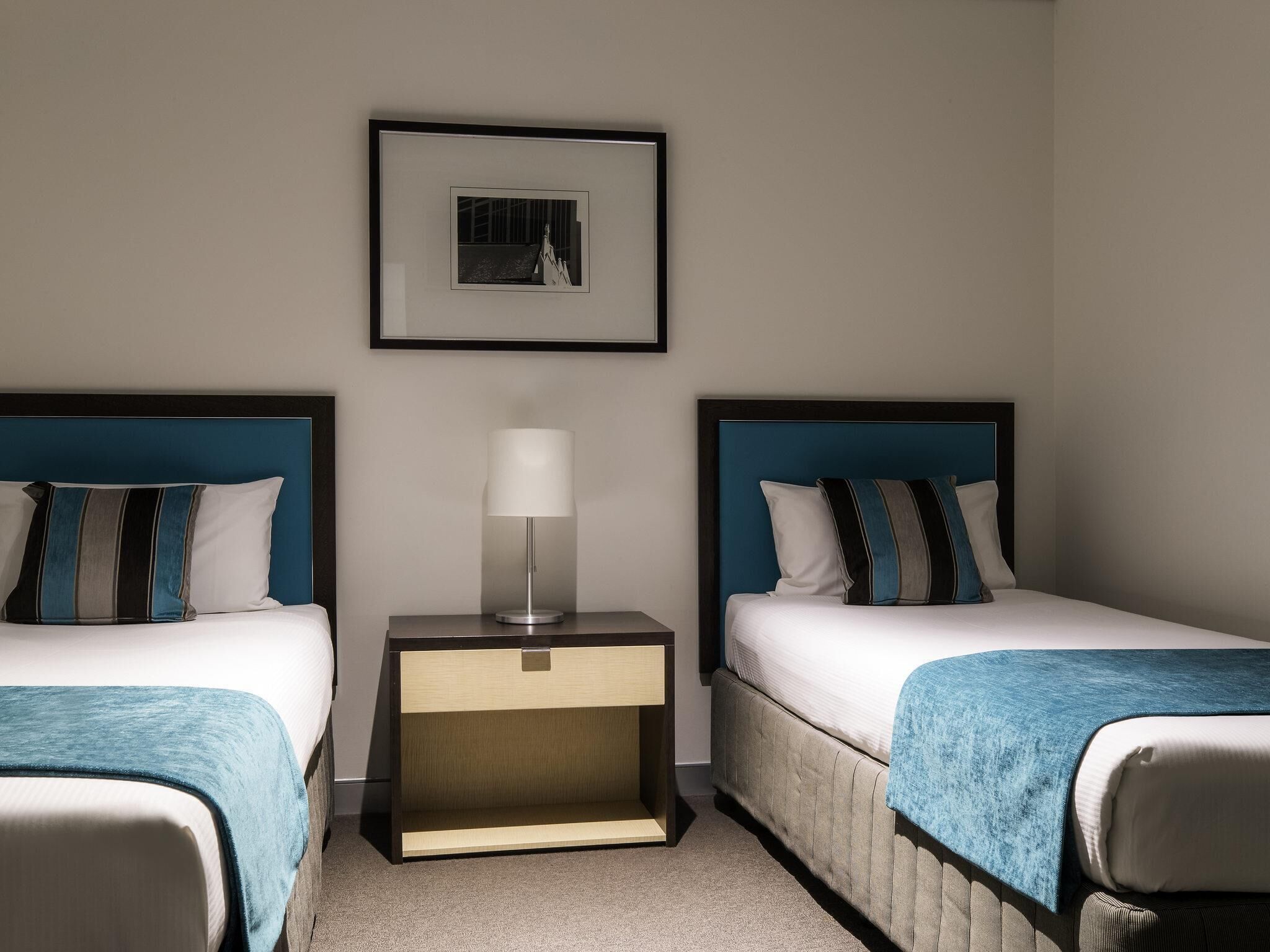 A hotel room with two beds, a nightstand with a lamp, and a framed picture on the wall.