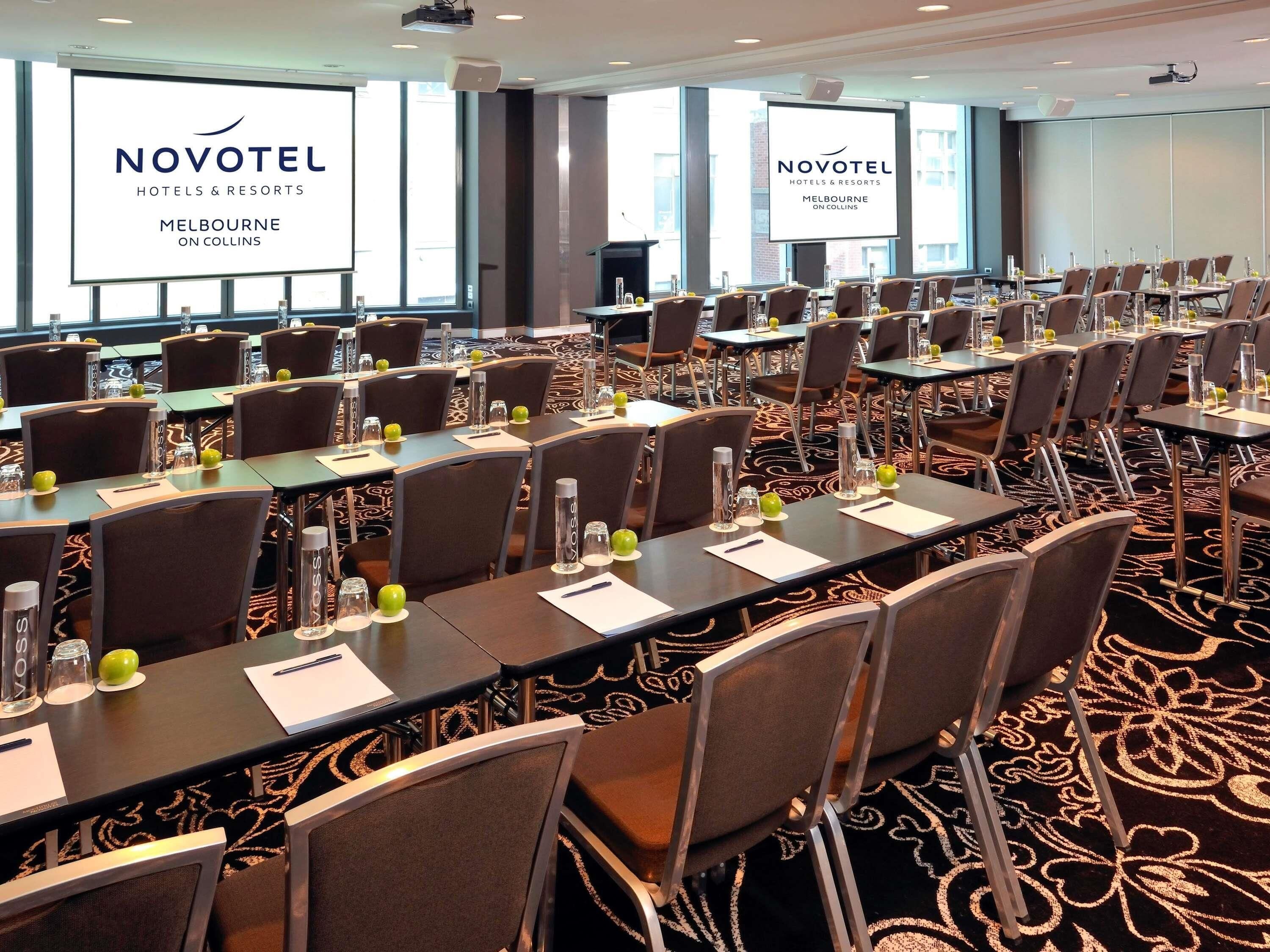 A conference room at Novotel Melbourne on Collins with multiple tables, chairs, and large windows.