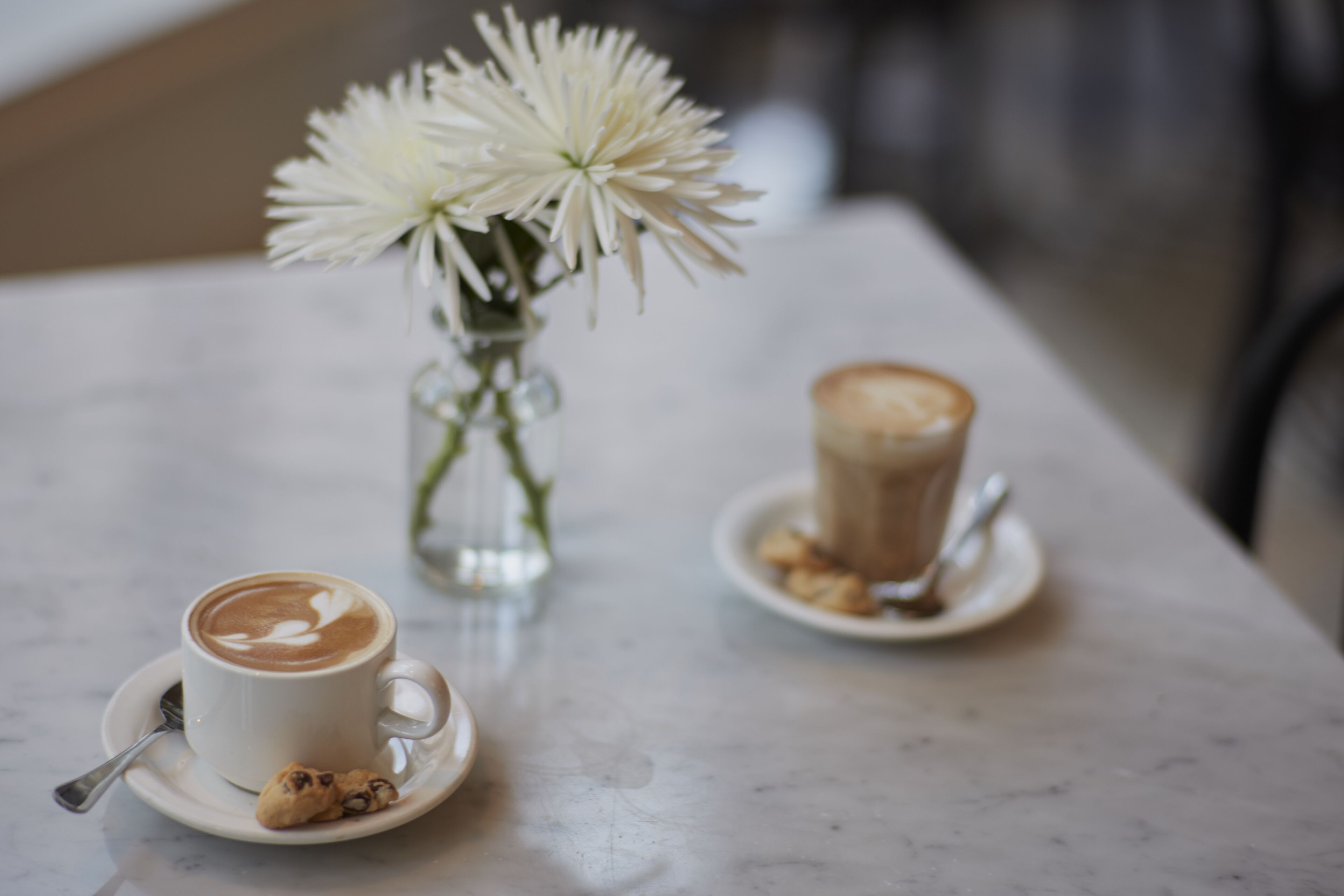 A table with a cup of coffee, a cookie, and a vase with flowers.