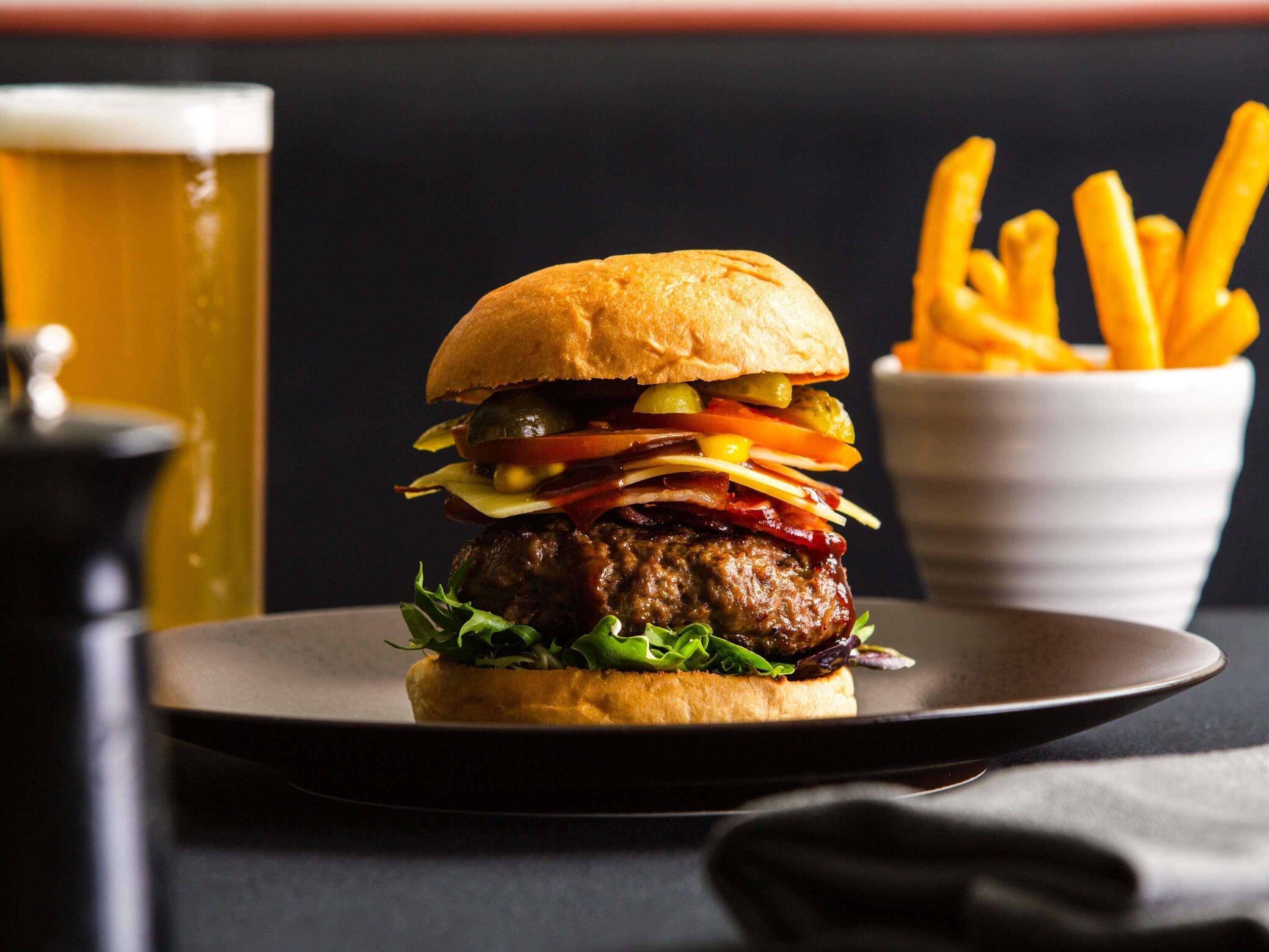 Ein Burger mit verschiedenen Belägen und ein Glas Bier auf einem Teller, dazu Pommes.