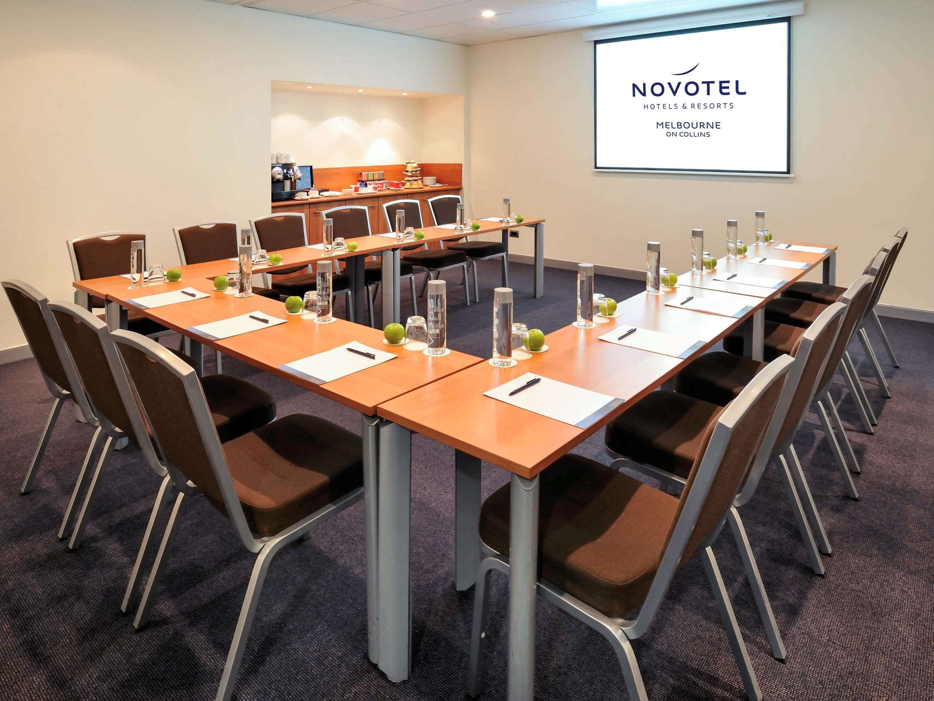 A conference room with a long table, chairs, and a projector screen displaying 'NOVOTEL HOTELS & RESORTS MELBOURNE ON COLLINS'.