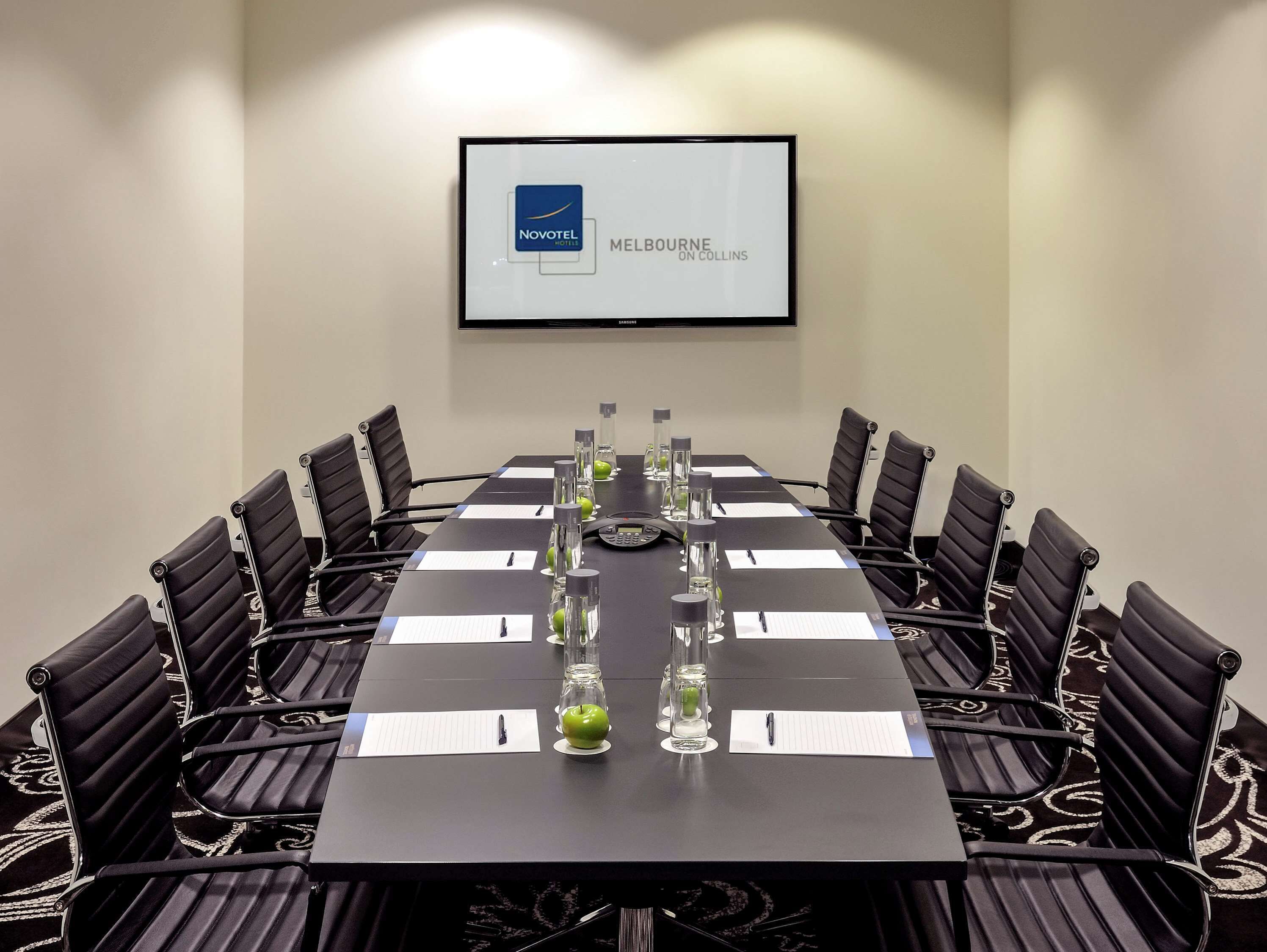 Ein Konferenzraum mit einem langen Tisch, schwarzen Stühlen und einer Projektionsleinwand, die 'Novotel Melbourne on Collins' zeigt.