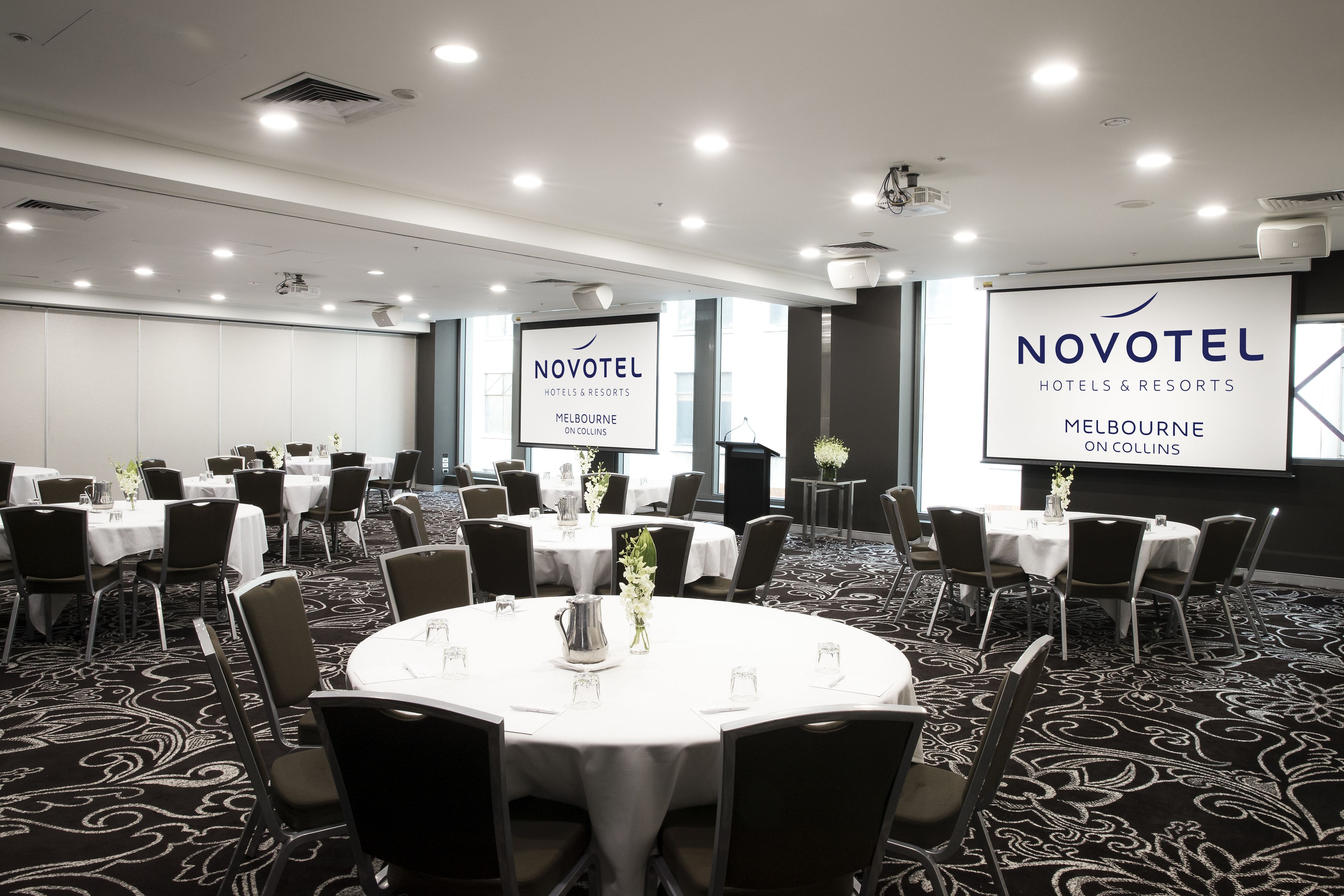 A conference room with round tables, chairs, and a projector screen displaying 'NOVOTEL HOTELS & RESORTS MELBOURNE ON COLLINS'.