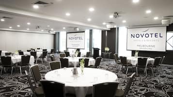 A conference room with round tables, chairs, and a projector screen displaying 'NOVOTEL HOTELS & RESORTS MELBOURNE ON COLLINS'.