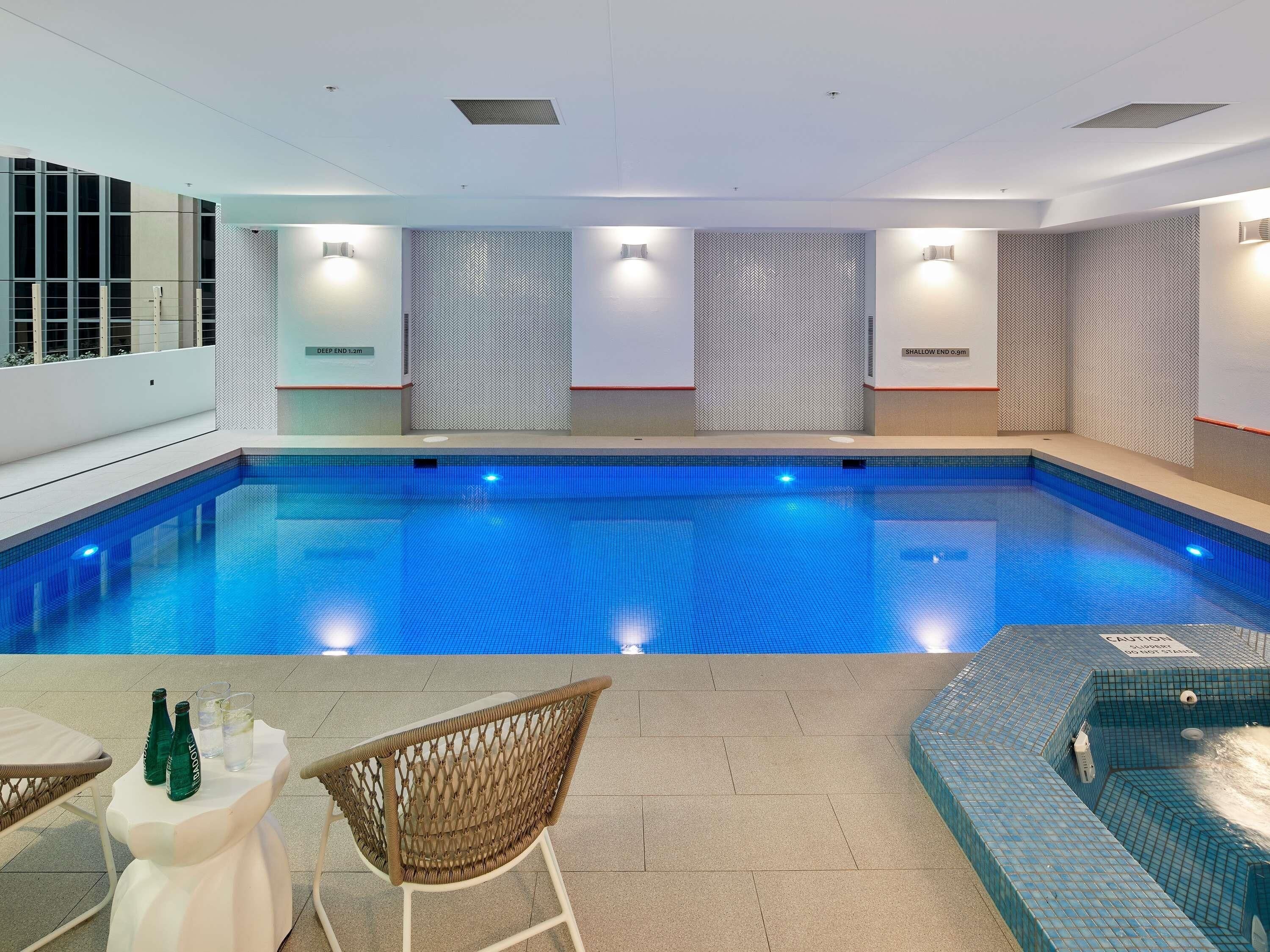 An indoor swimming pool with a tiled floor, a lounge area with chairs and a table, and a jacuzzi area.