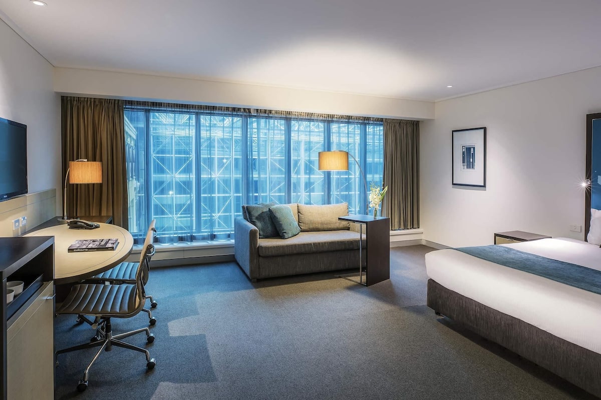Habitación de hotel con cama, escritorio con silla, sofá, televisor y ventanas grandes con cortinas.
