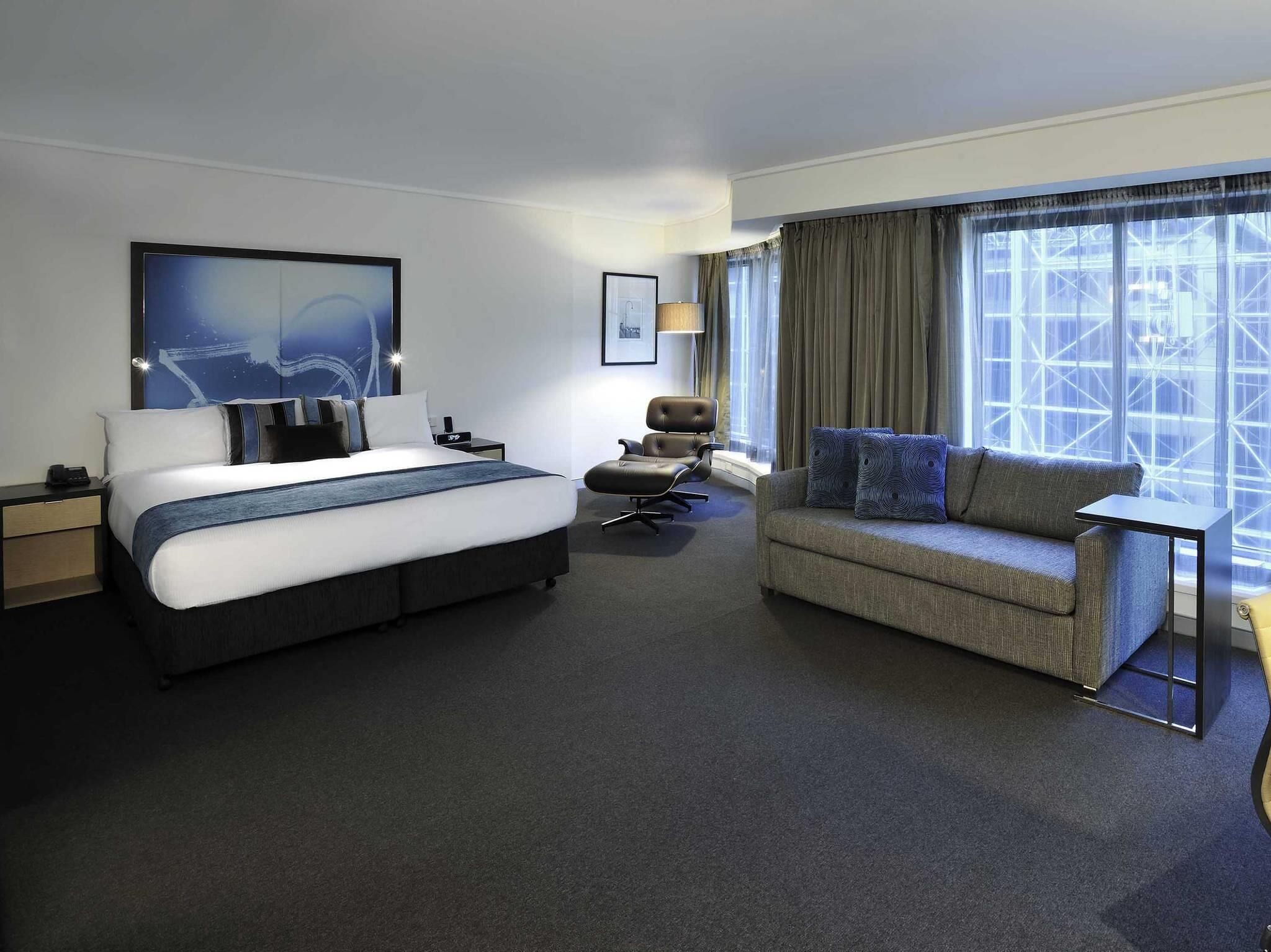 A hotel room with a bed, a sofa, a chair, a desk, and a large window with curtains.