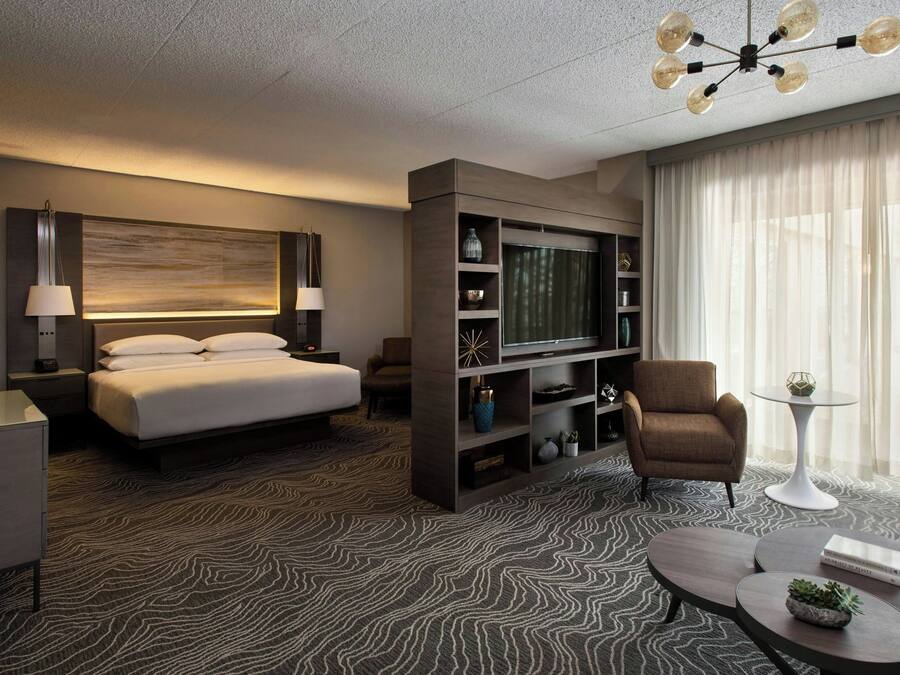 A modern hotel room with a large bed, a TV, a seating area, and a chandelier.