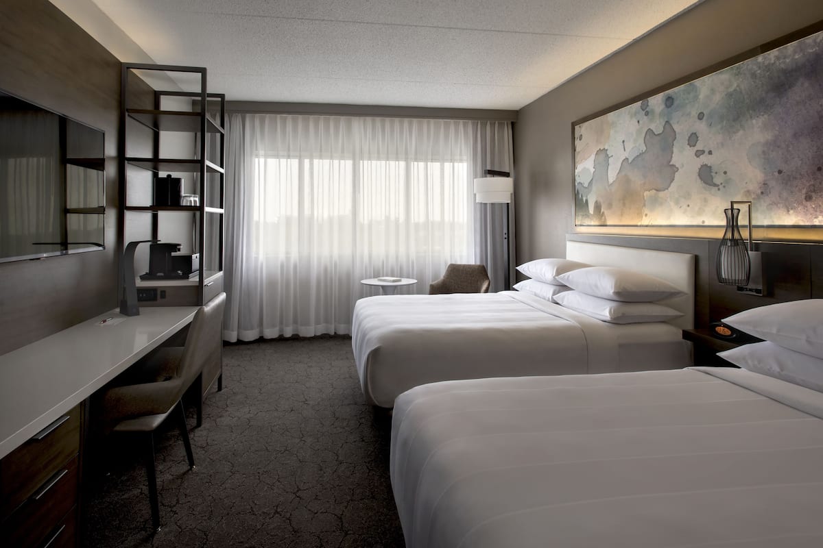Une chambre d’hôtel avec deux lits, un bureau, une chaise, une petite table ronde, une lampe et un grand tableau abstrait accroché au mur.