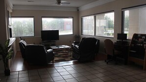 A living room with a television, two armchairs, a coffee table, and a desk with a computer. There are large windows with blinds, a ceiling fan, and a tiled floor. - Deerfield Inn & Suites (Madison)