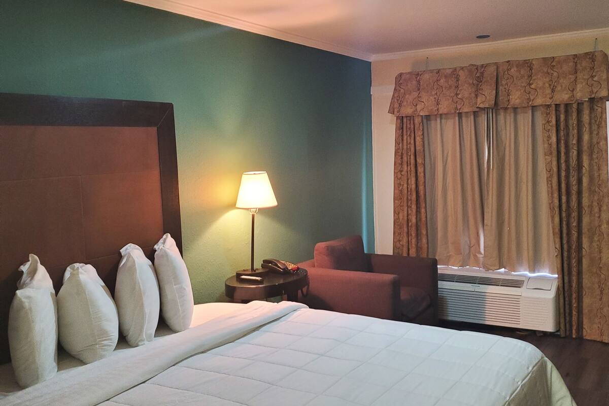 A hotel room with a large bed, a desk with a lamp, a chair, and an air conditioning unit.