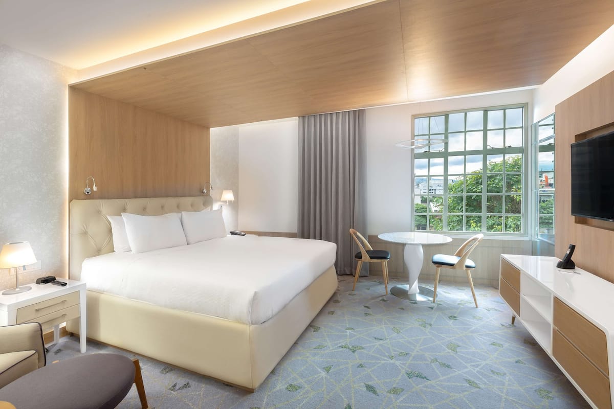A modern hotel room with a large bed, a desk, a chair, a small table, and a TV.