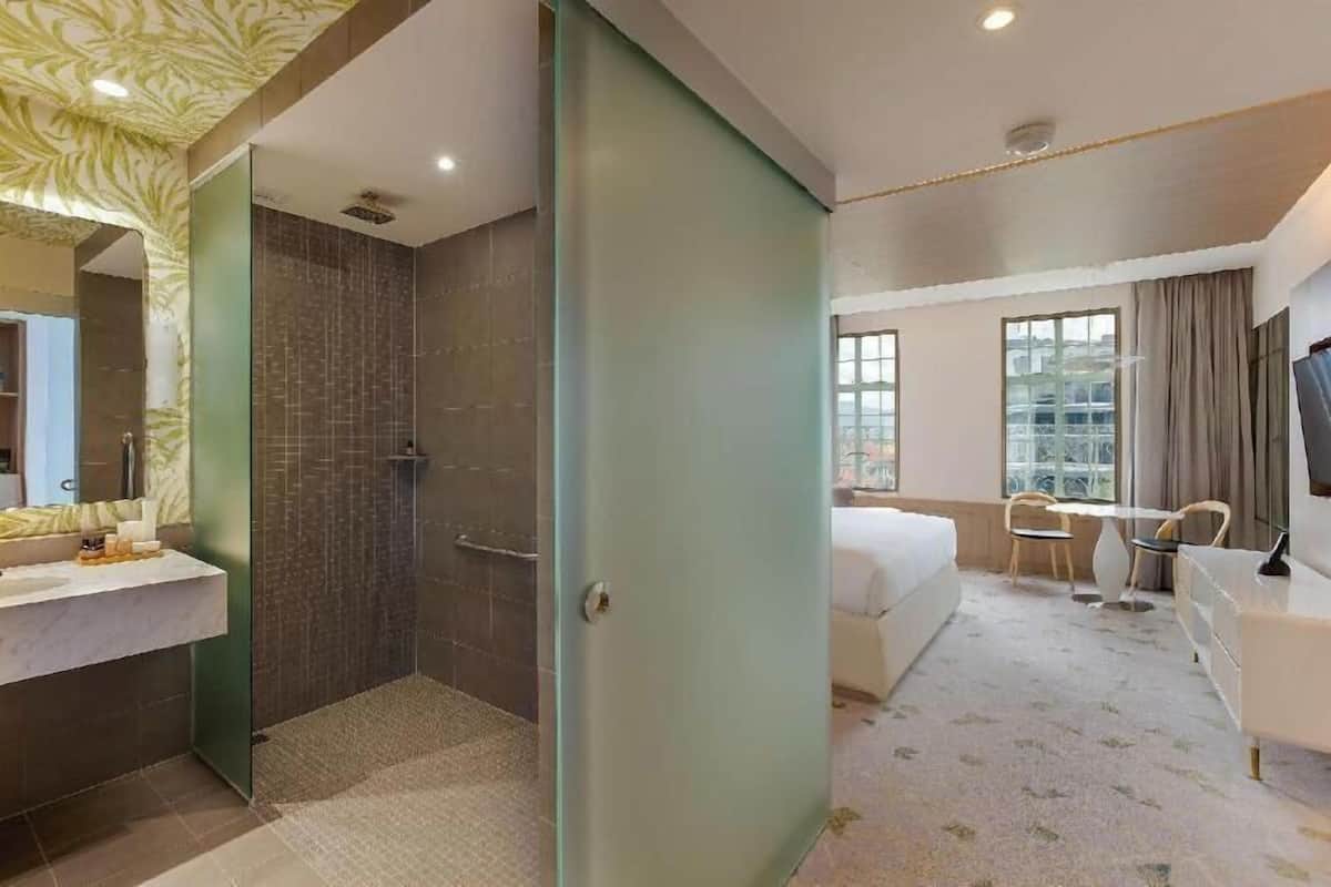 A hotel room with a shower, a bed, a desk, a chair, and a window.