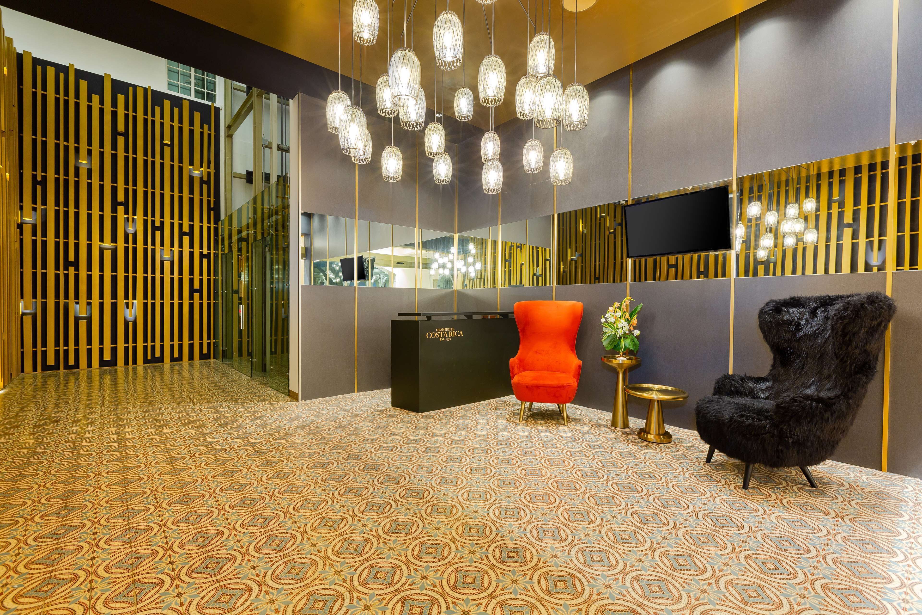 A reception area with patterned carpet, a black reception desk, a red chair, a black chair, and hanging crystal chandeliers.