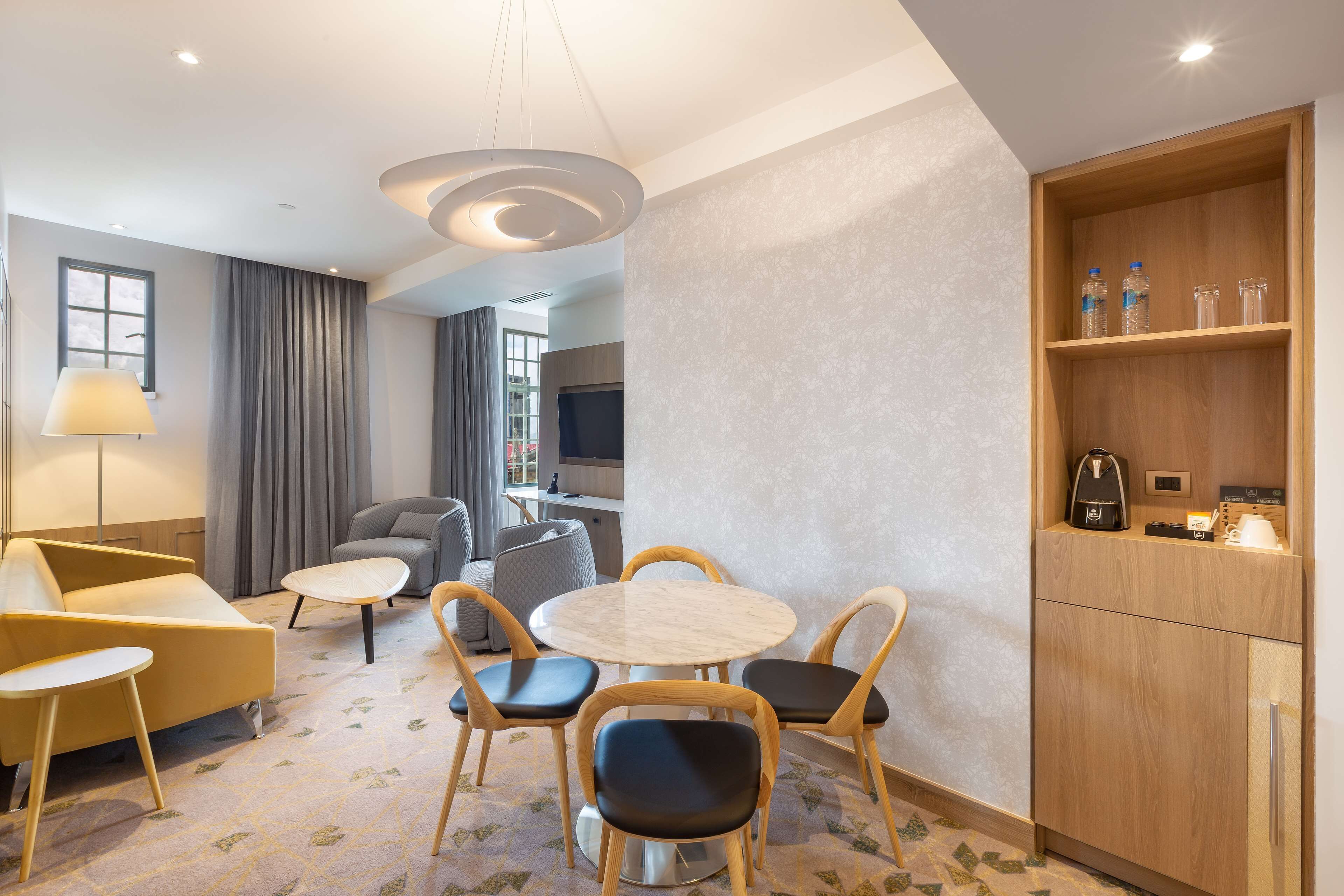 A modern hotel room with a round dining table, four chairs, a sofa, a lamp, a TV, and a minibar.