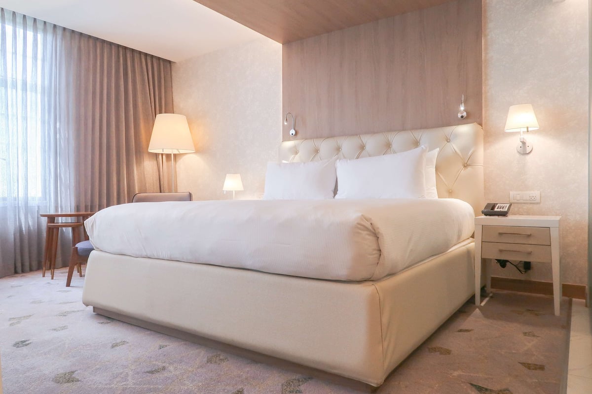 A hotel room with a large bed, a bedside table with a lamp, a chair, and a desk.